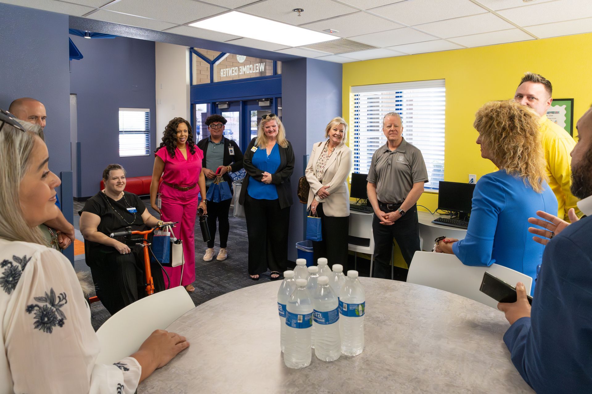 People standing in a meeting room around a table with water bottles, listening to a discussion.