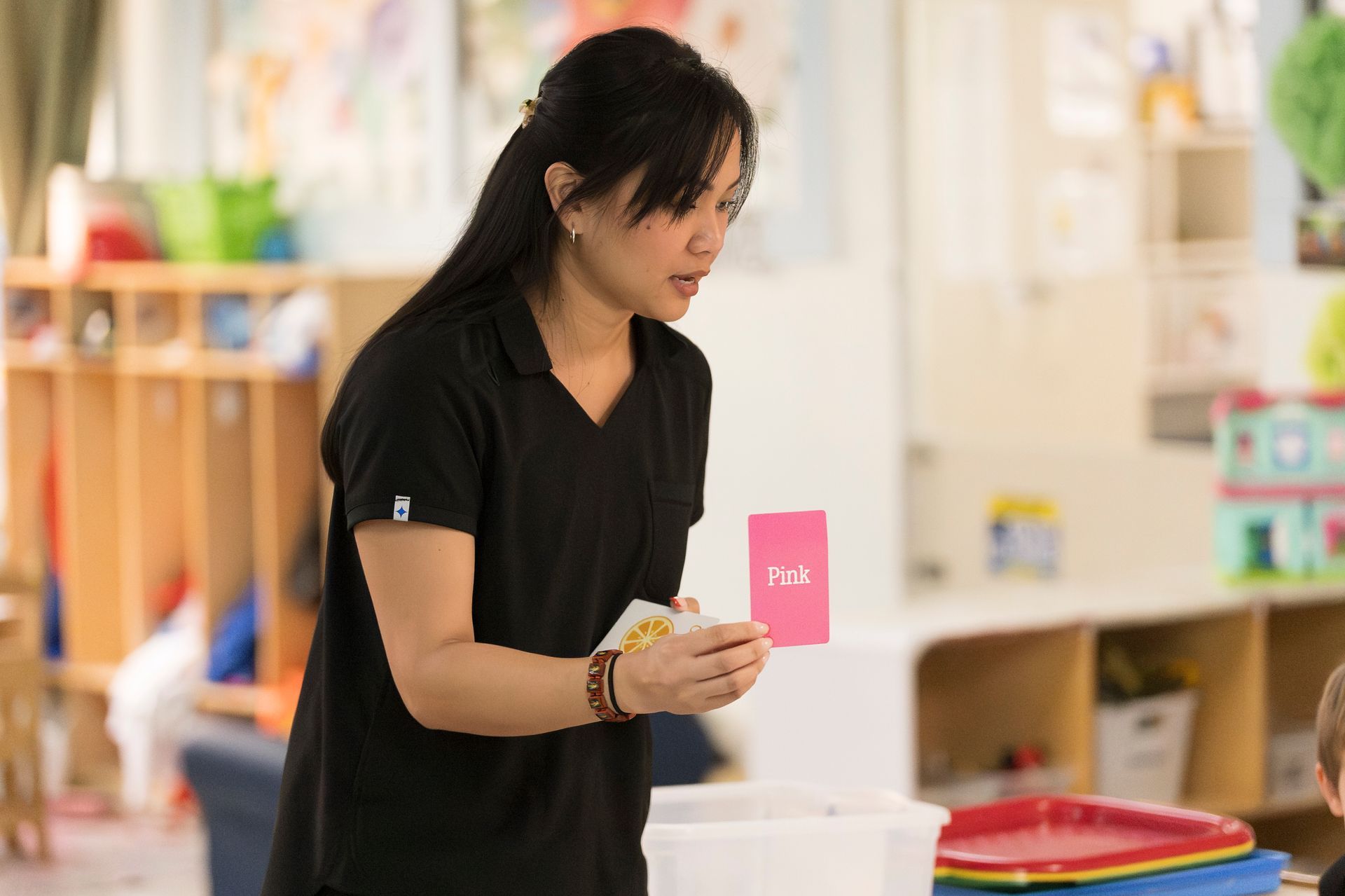 Woman in black shirt holding a pink card in a classroom, leaning over a table.