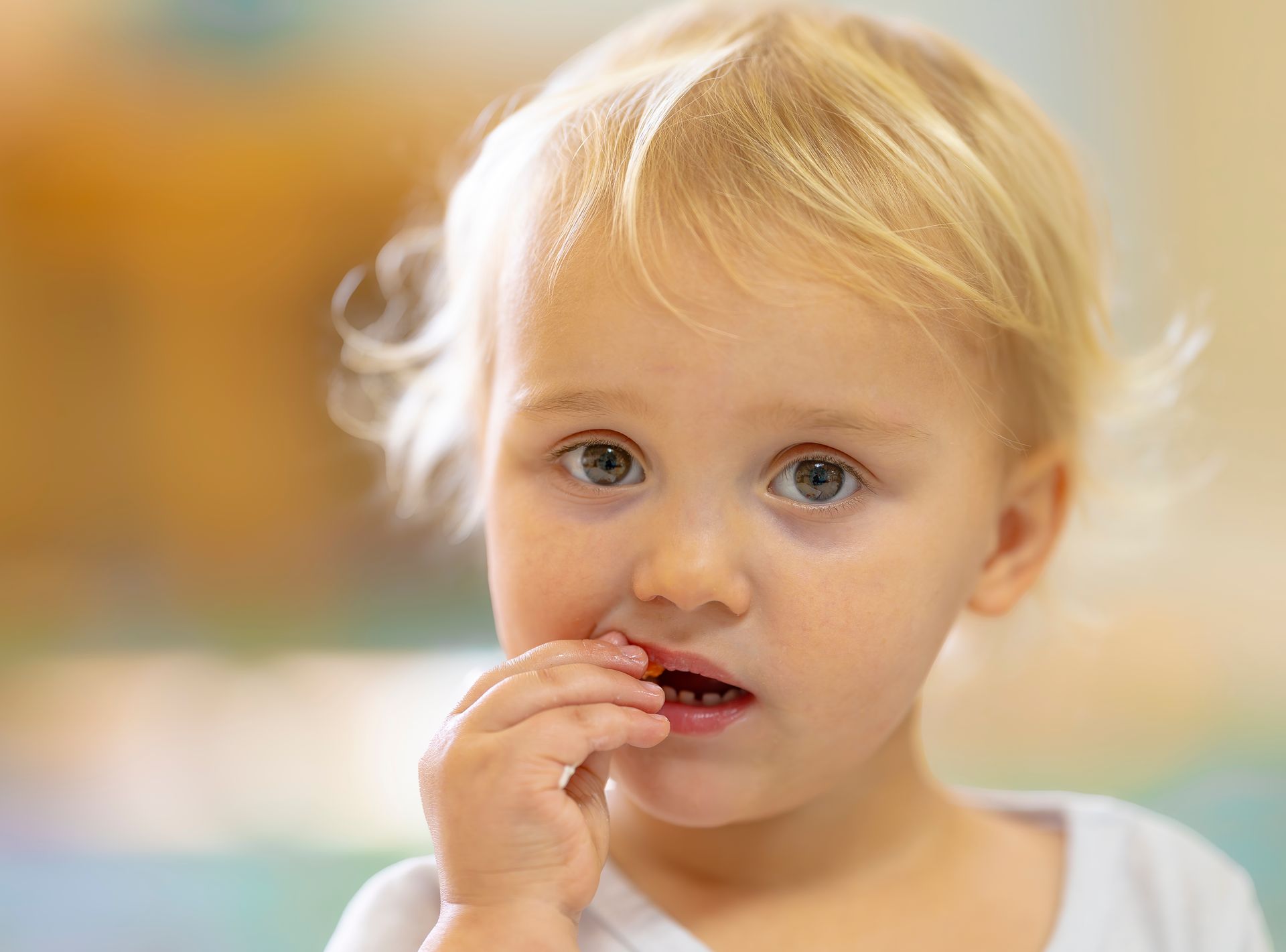 Child with blonde hair touching lips, looking at the camera against a soft blurred background