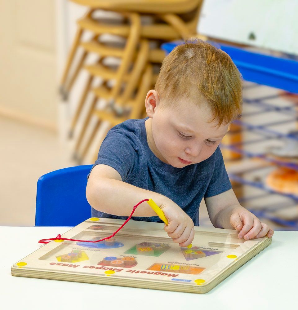 Child playing with a wooden activity board at a table, concentrating on moving pieces with a wire loop