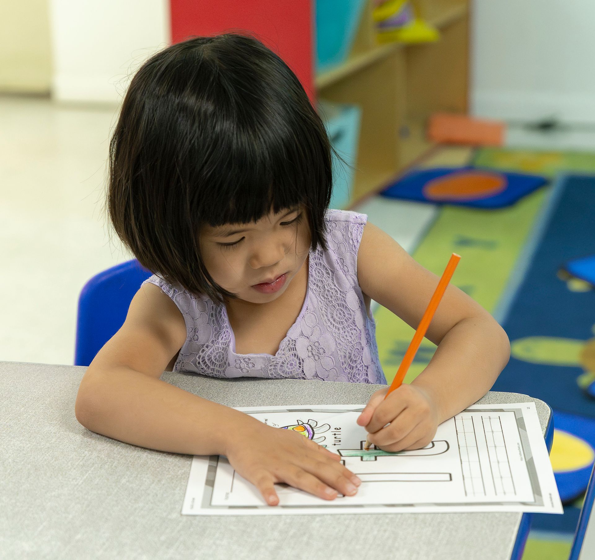 Child coloring at a table with a pencil, leaning over a worksheet in a classroom.