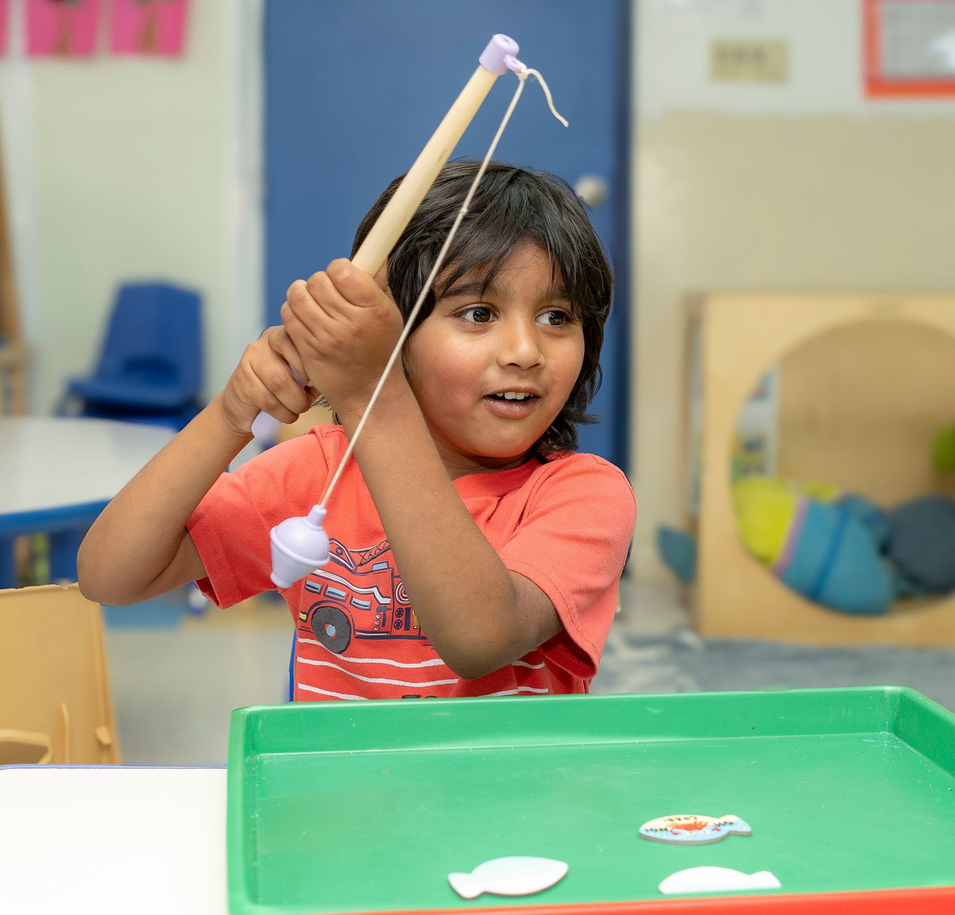 Child at a table using a fishing toy, smiling and holding a wooden rod in a classroom.