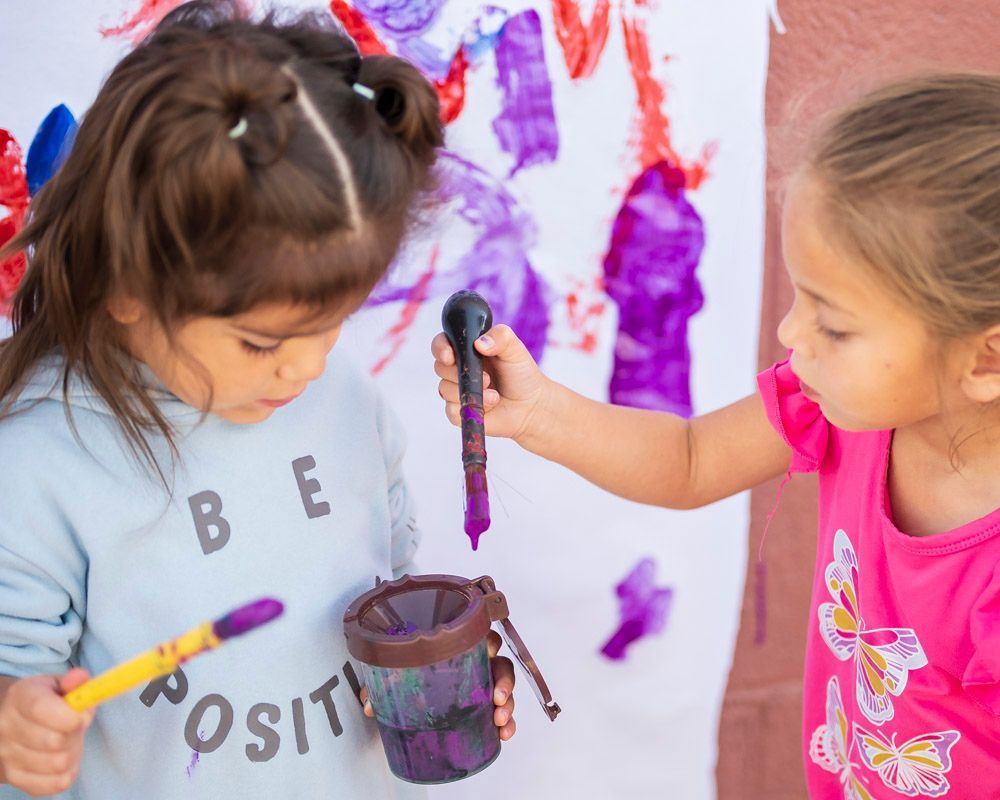 Two children painting a wall, one holding a paintbrush and bucket of purple paint