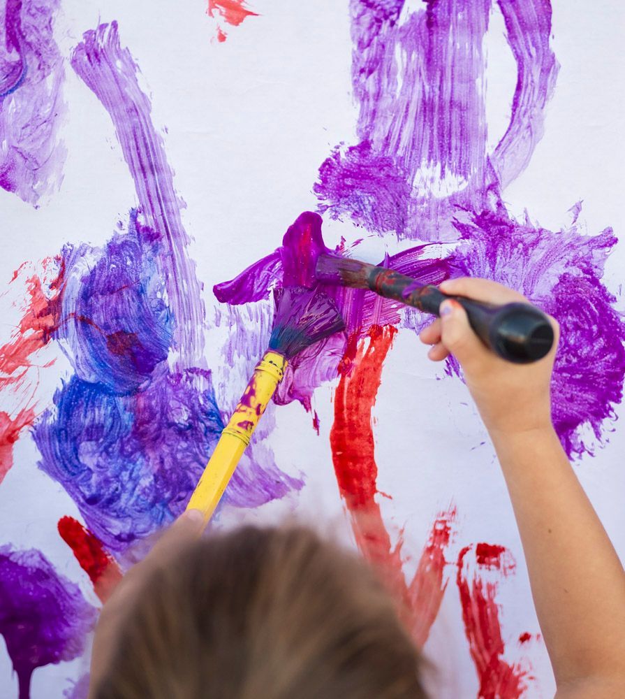 Child painting colorful purple, red, and blue brushstrokes on a white wall with a paintbrush
