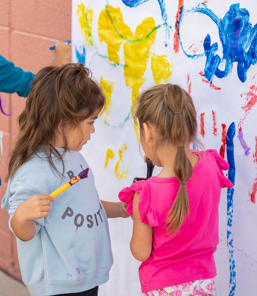 Two children painting colorful shapes on a large wall mural with brushes