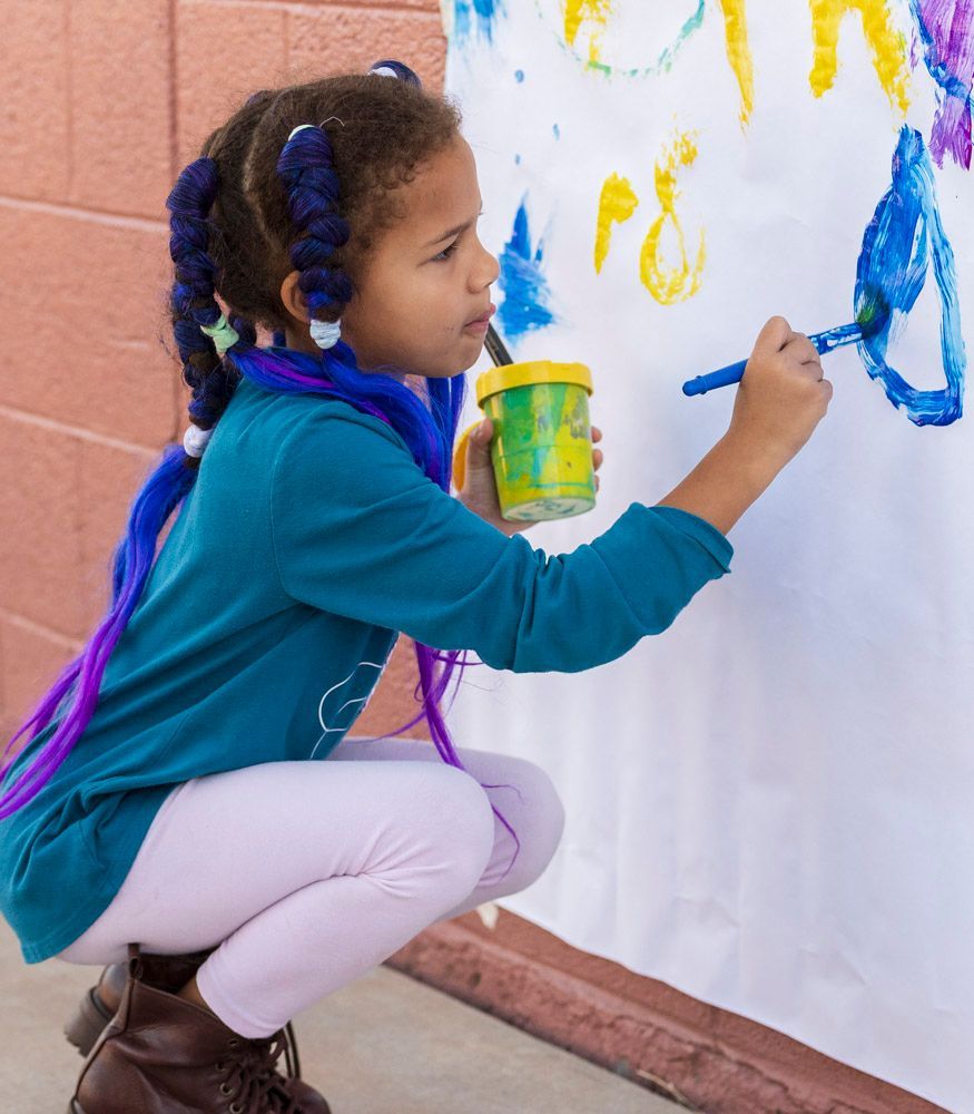 Child crouching and painting a colorful mural on a wall with a small paint cup in hand