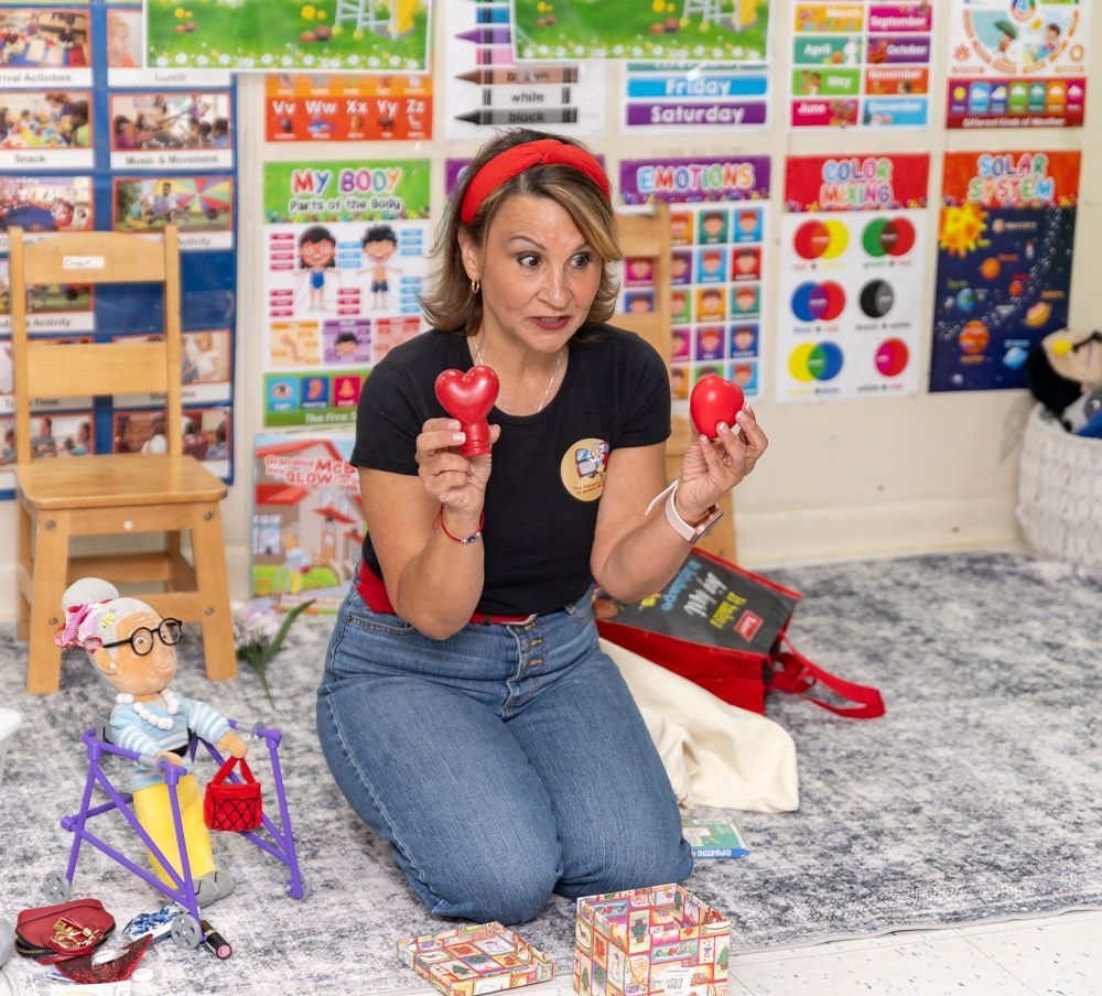Woman kneeling in classroom, holding two red toys, with colorful learning posters and toys around her.