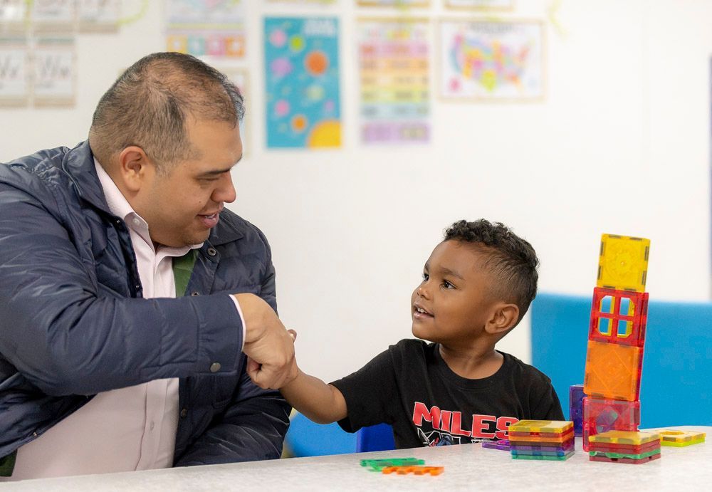 Man and child smiling and bumping fists at a colorful table with toys in a classroom