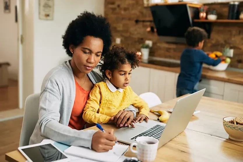 Adult and child working on a laptop at a kitchen table, with another child in the background