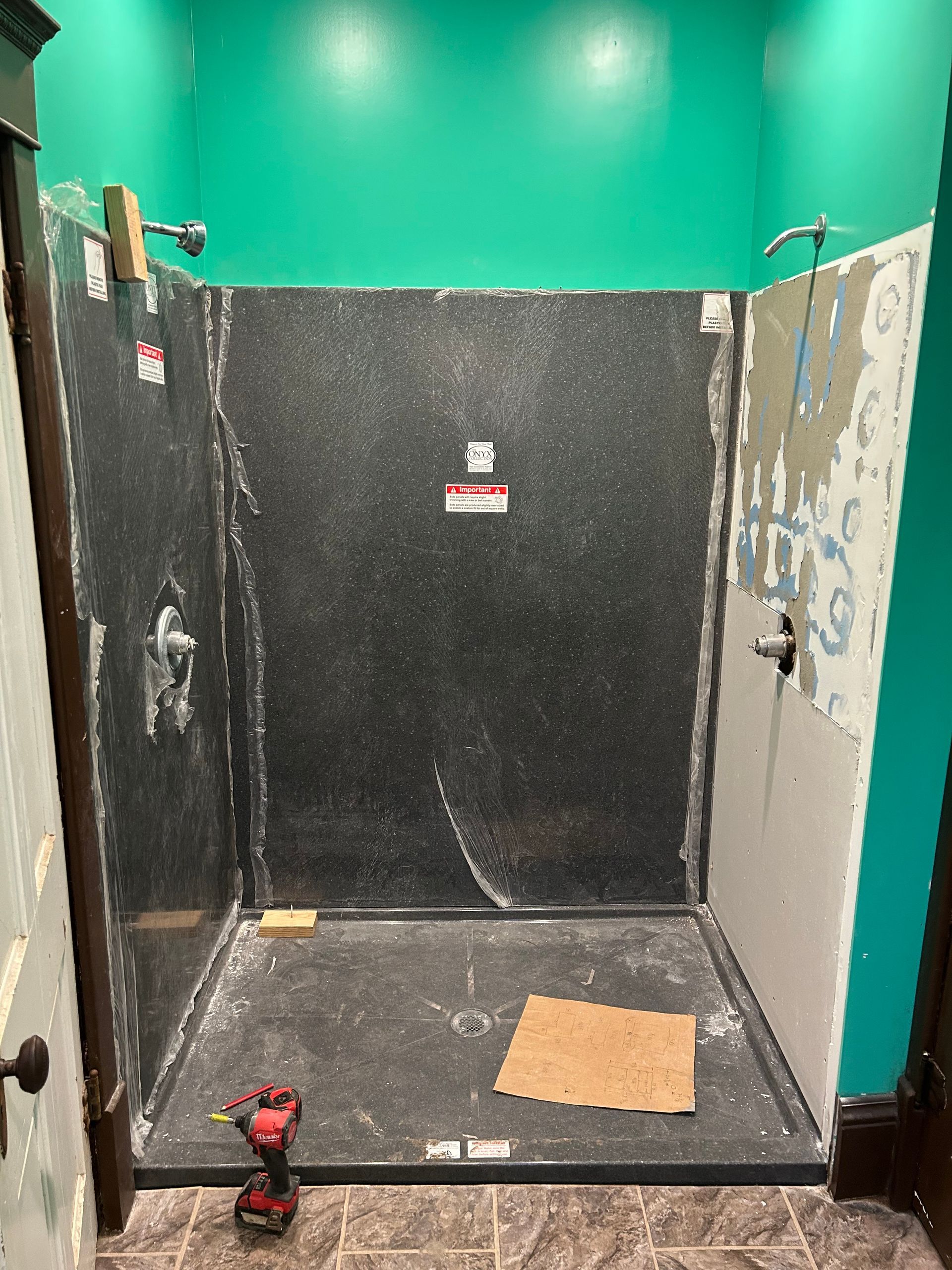 Small bathroom stall with dark walls, teal upper wall, red drill on the floor, and a tan square tile.