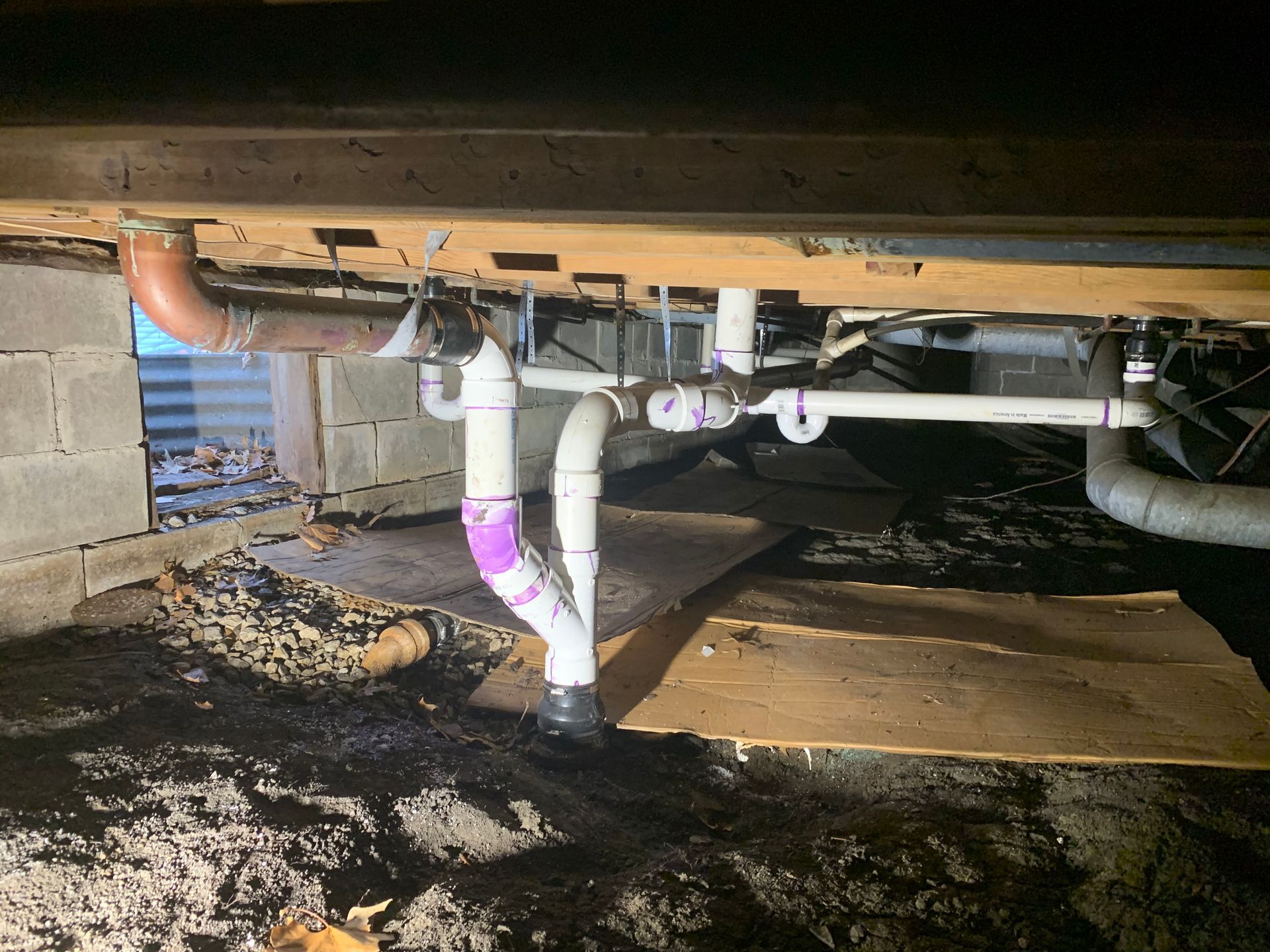 Exposed plumbing under a house, with white and copper pipes and purple primer on joints.