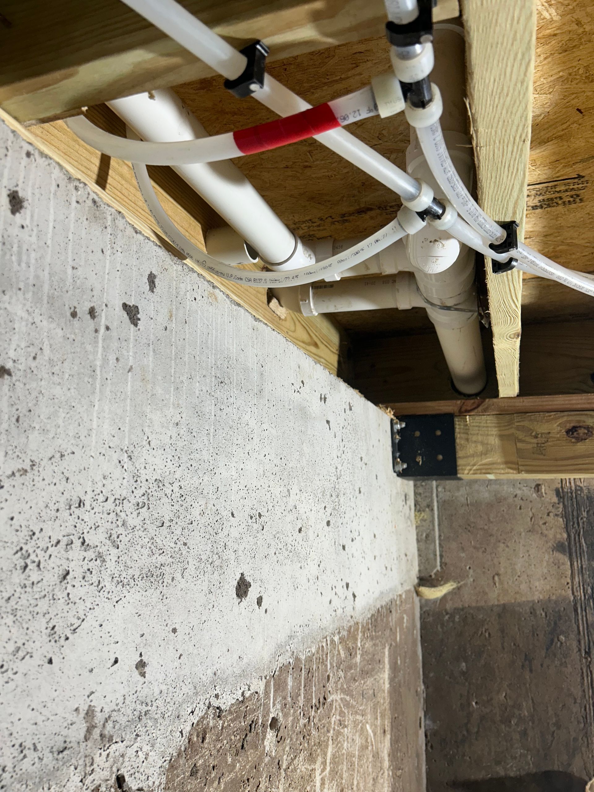 Exposed white pipes under a wooden floor joist with a red clamp and concrete wall nearby