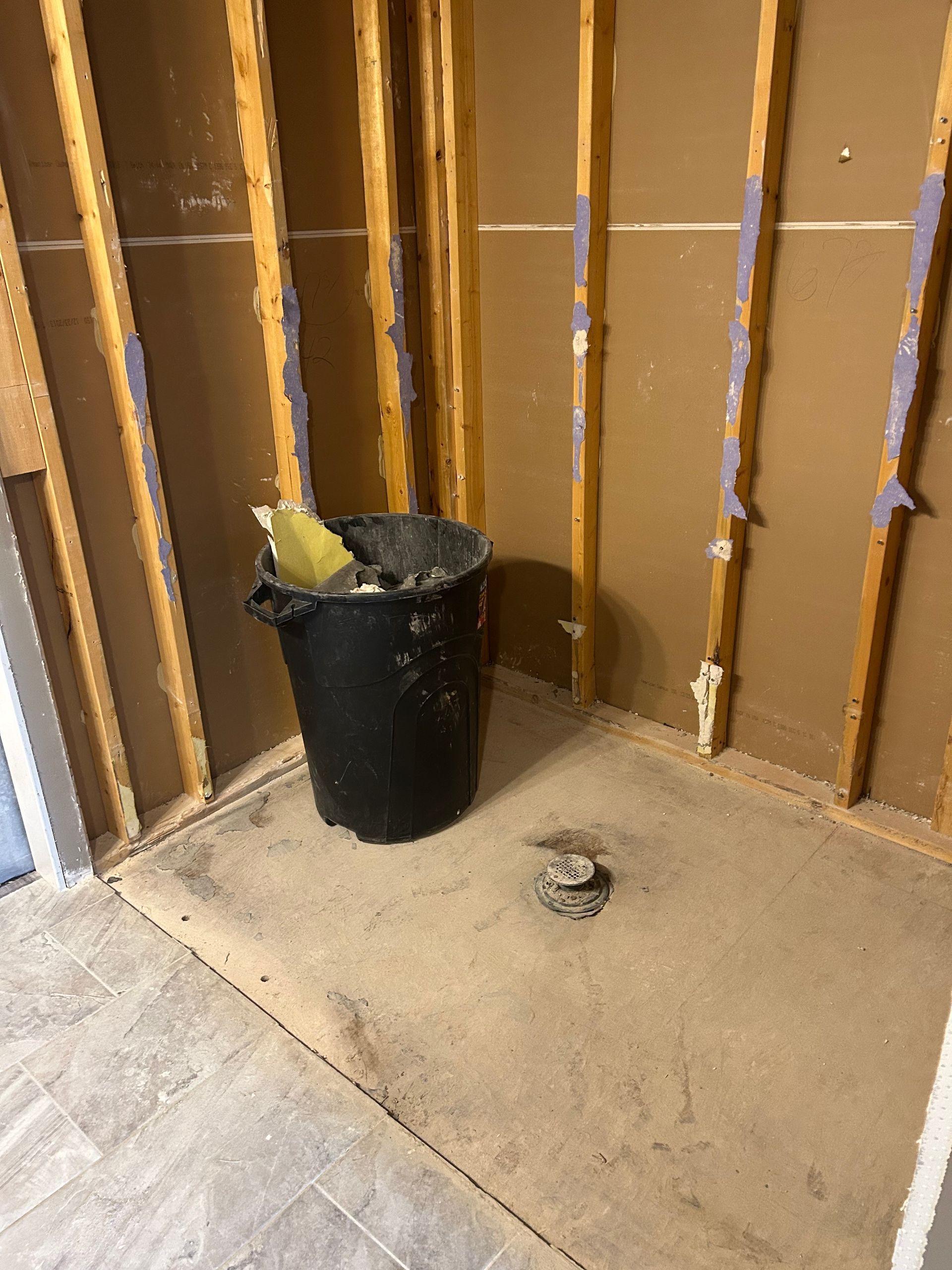 Partially finished room corner with exposed studs, black trash can, and a drain on the floor