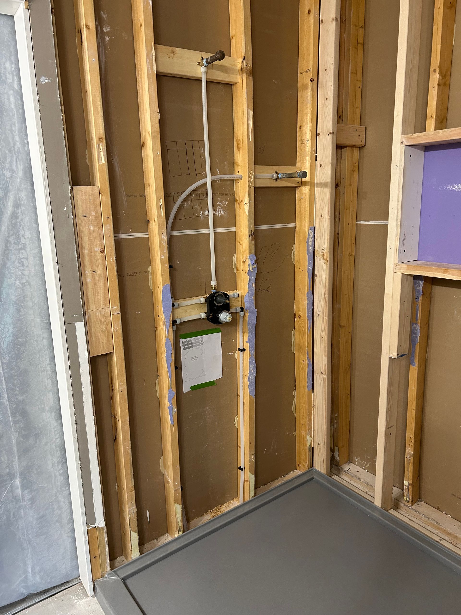 Unfinished interior wall framing with exposed studs and plumbing beside a gray shower pan base.