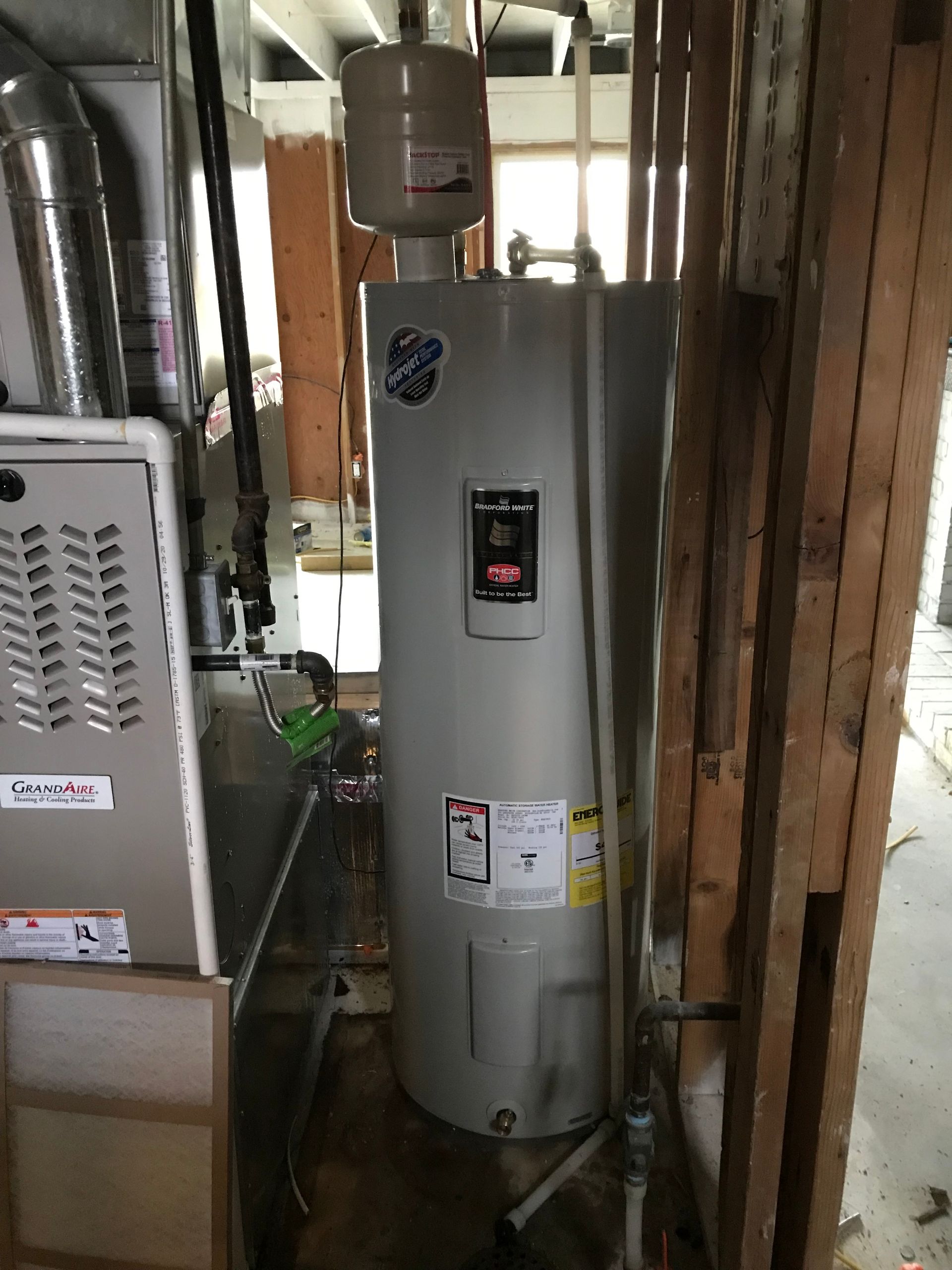 Tall gray water heater in a narrow utility room with pipes and ducting around it.