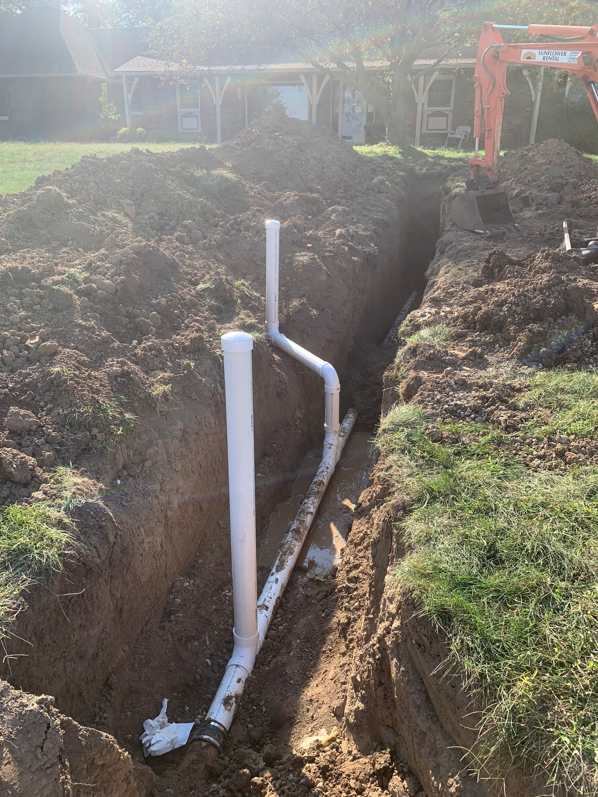 Trenched yard with white PVC pipes running through a narrow ditch near a house under construction.