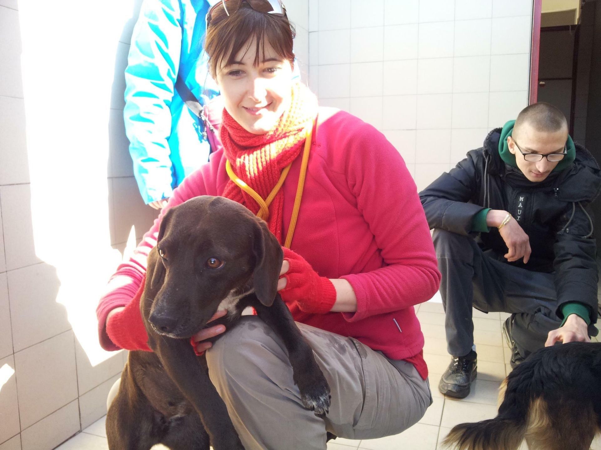 Due persone sono accovacciate in un ambiente chiuso con due cani, uno nero davanti e un altro parzialmente visibile sulla destra.