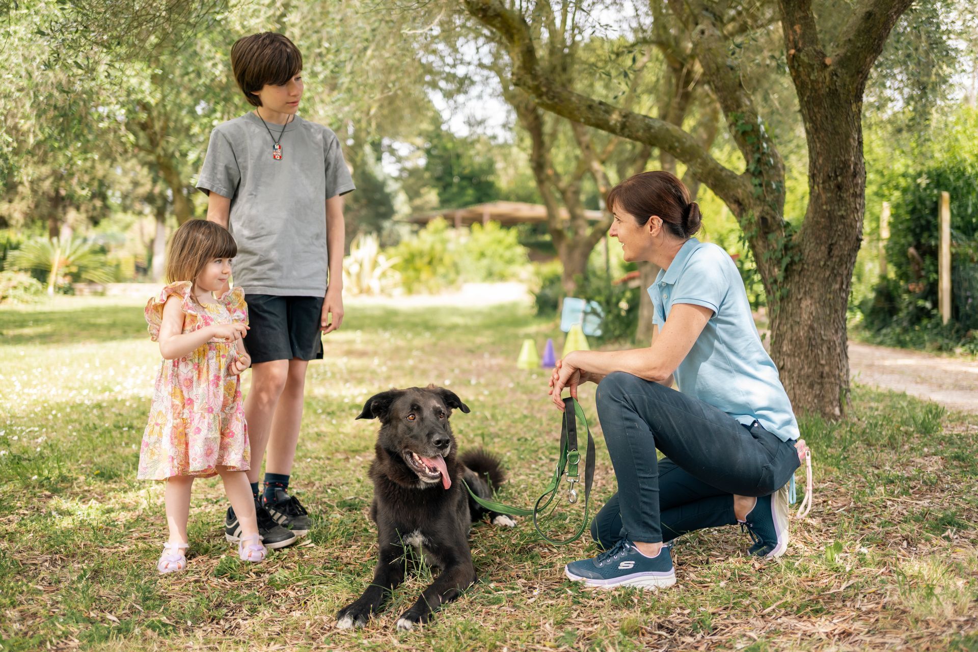In un parco soleggiato, una donna è inginocchiata accanto a un cane nero, mentre un uomo e un bambino piccolo le stanno vicino.