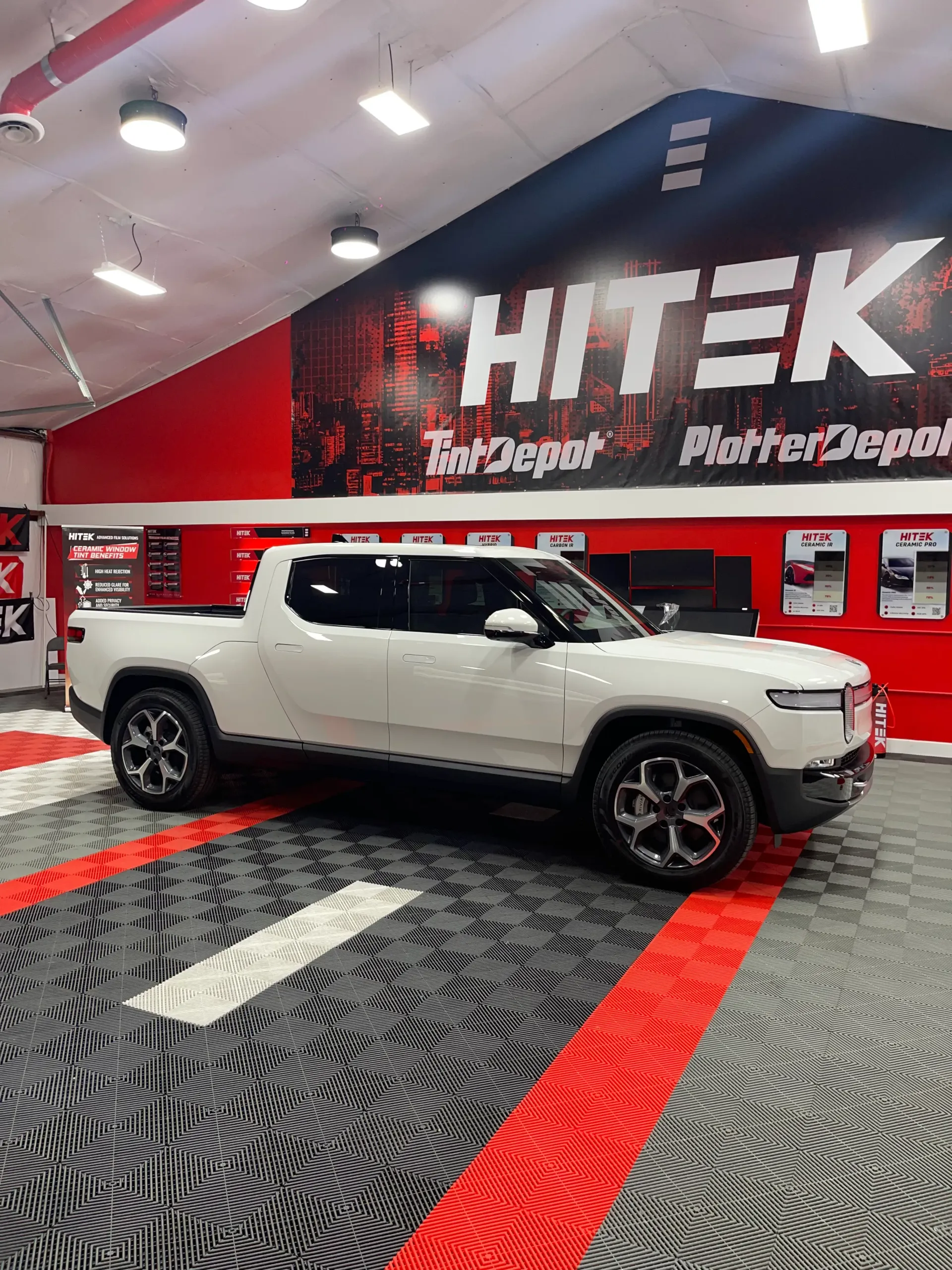 White Rivian pickup truck inside a car repair shop with a red and black logo and checkered floor.