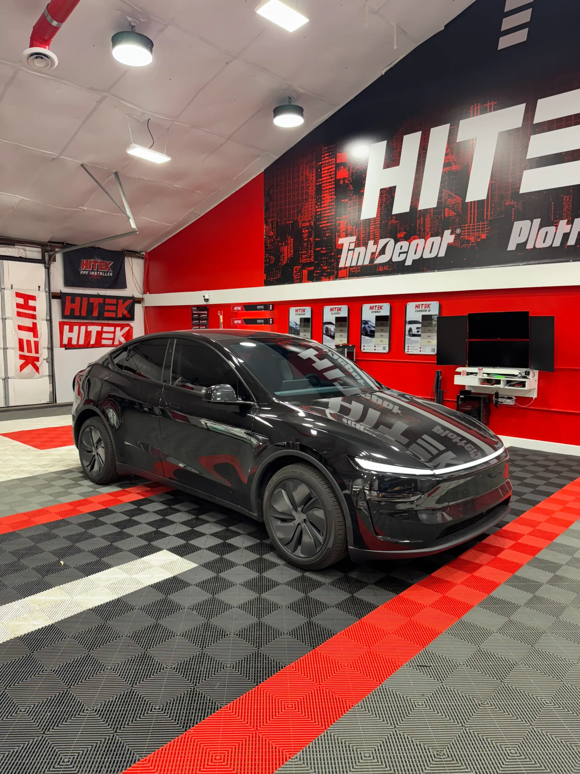 Black electric car in a shop with red accents. HITEK Tint Depot sign in background.