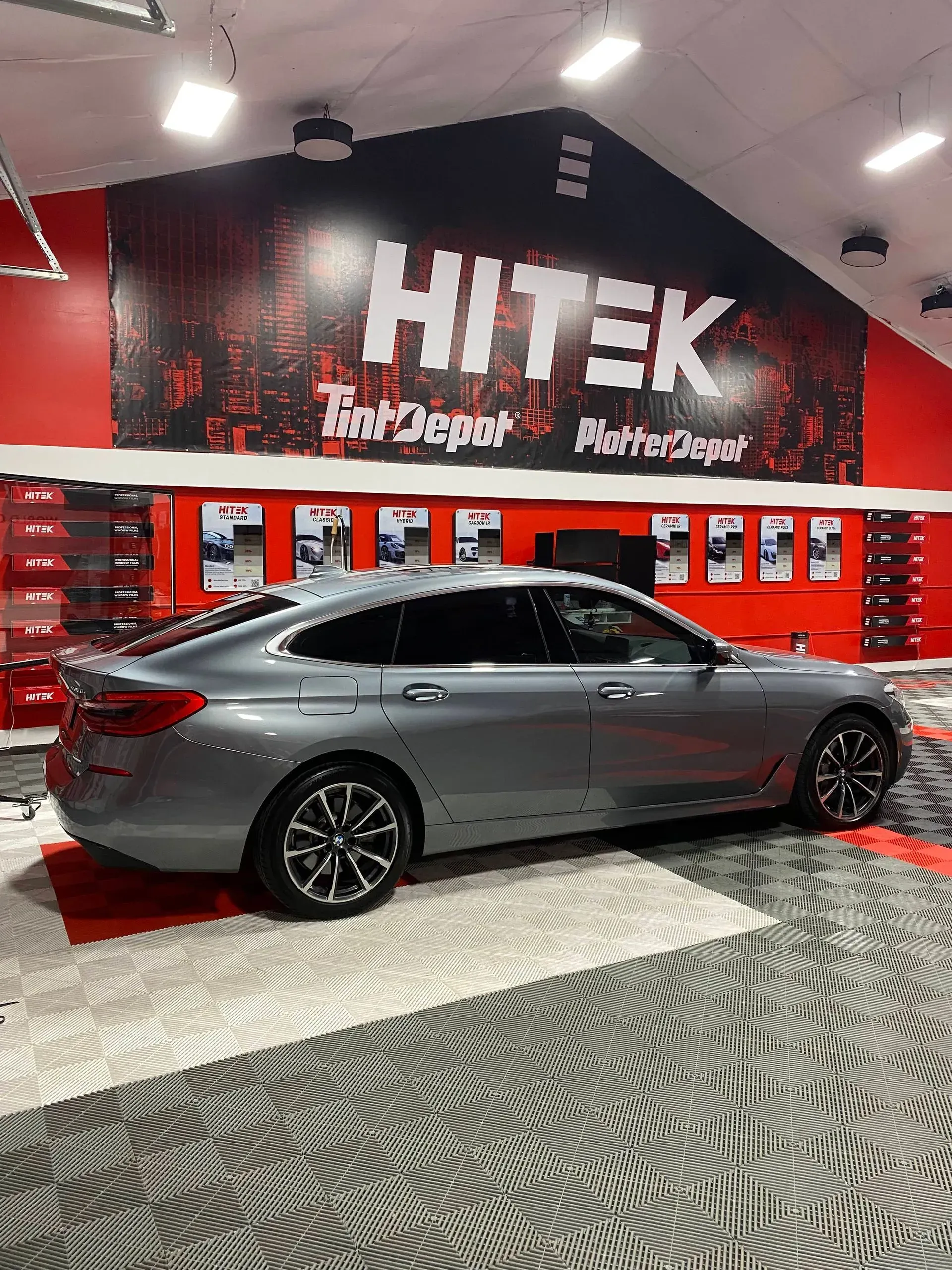Gray BMW sedan parked inside a HiTek shop; red and black logo in background.