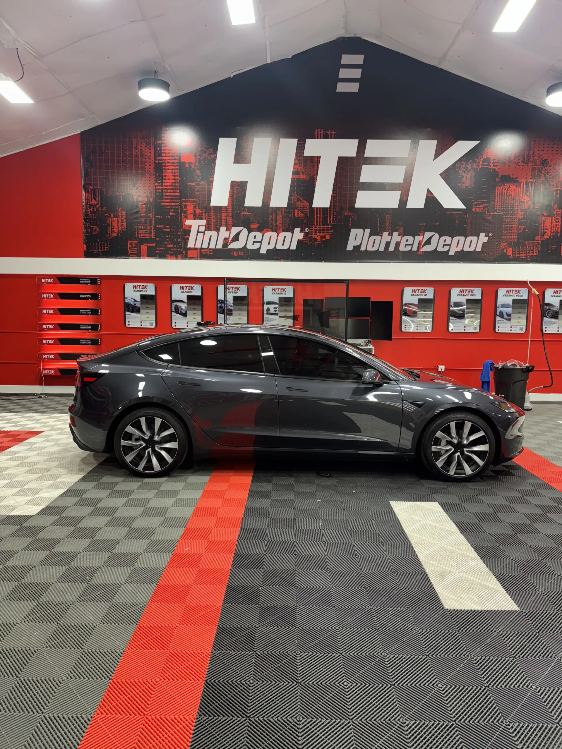 Gray Tesla sedan inside a HiTek Tint Depot shop; red, white, and gray floor tiles; red and black wall graphics.
