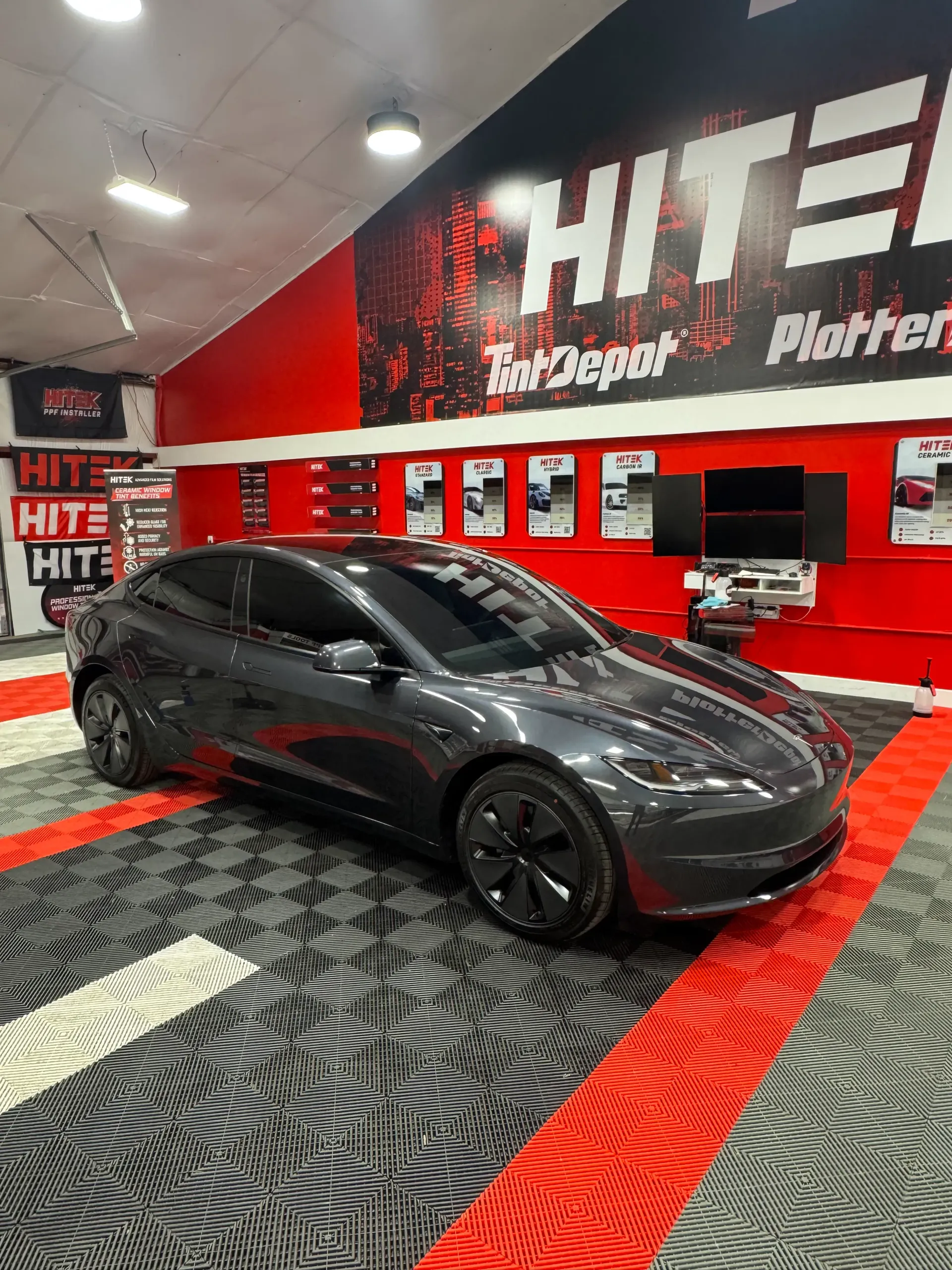 Dark gray Tesla in a red and black shop, possibly a tinting service, with a patterned floor.