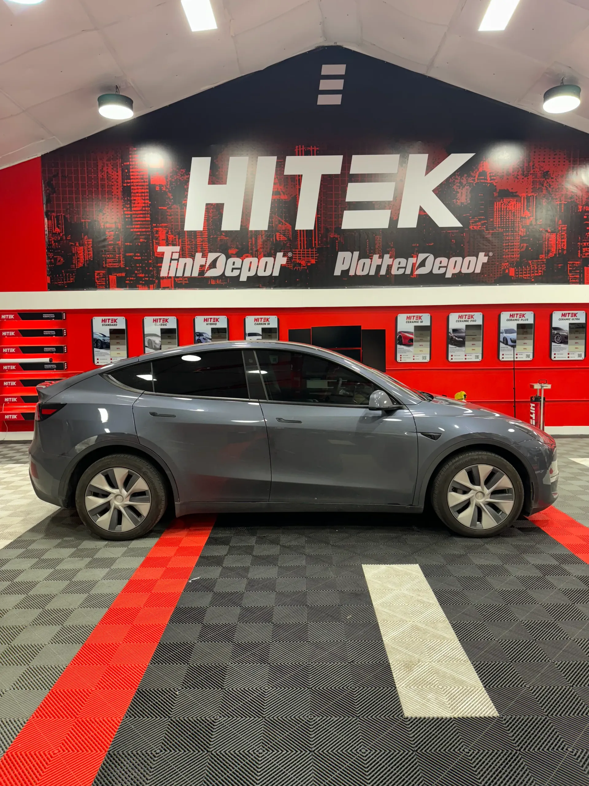 Gray Tesla Model Y car in a tint shop, with the shop's logo and red/black flooring.