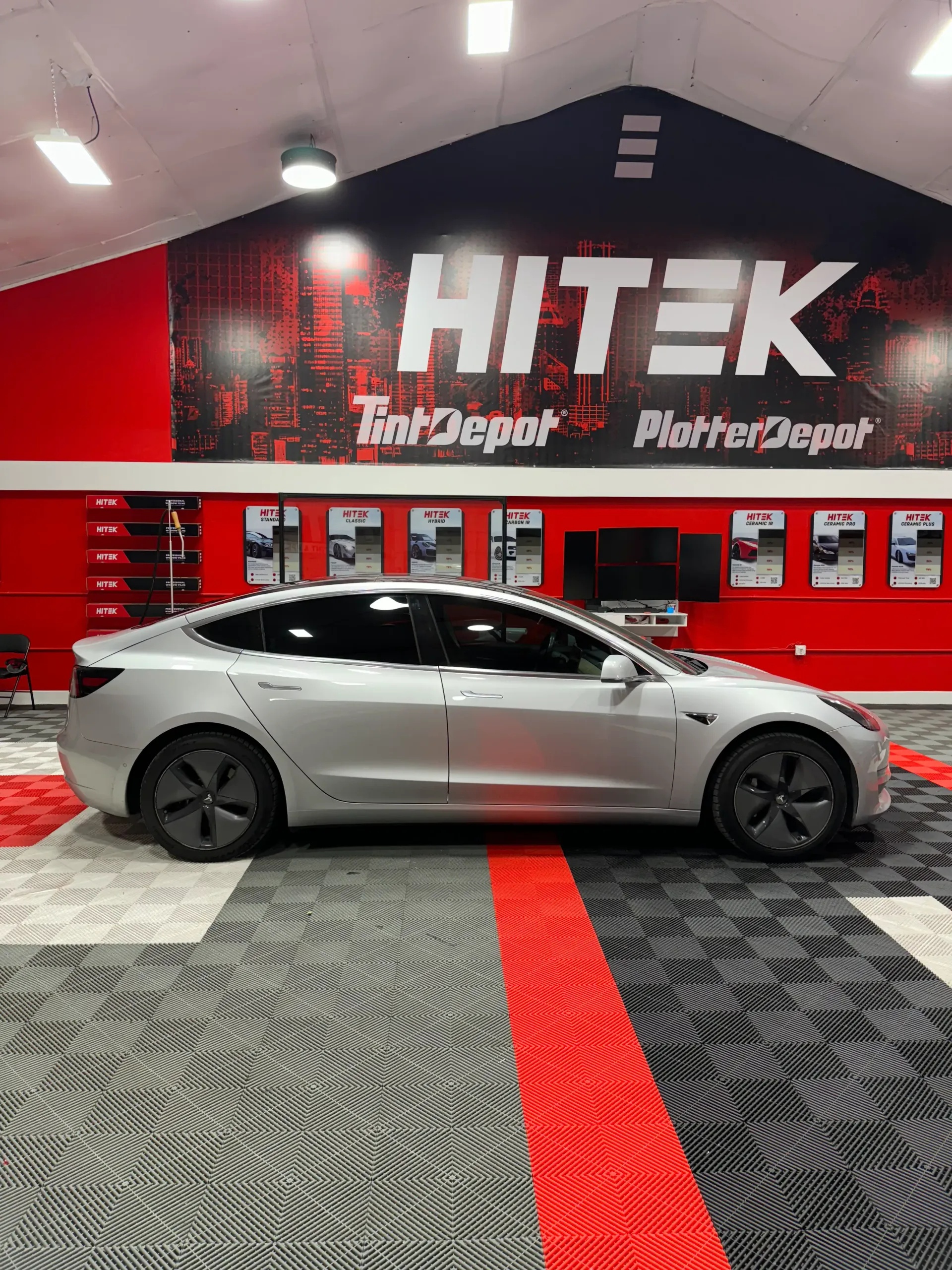 Silver Tesla sedan in a shop with red and black accents, under a Hitek sign.
