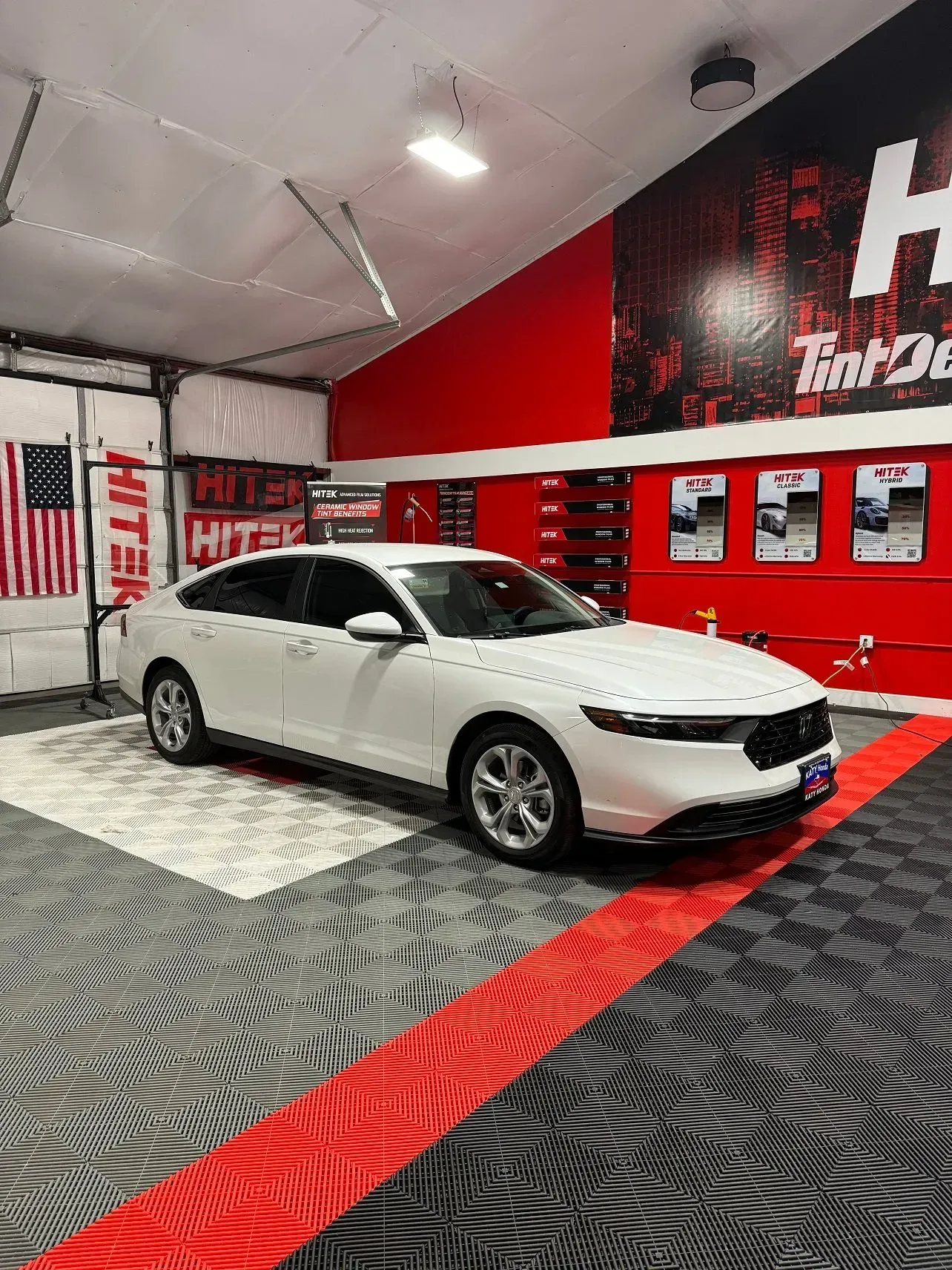White sedan in a garage, next to an American flag and a red wall with graphics.
