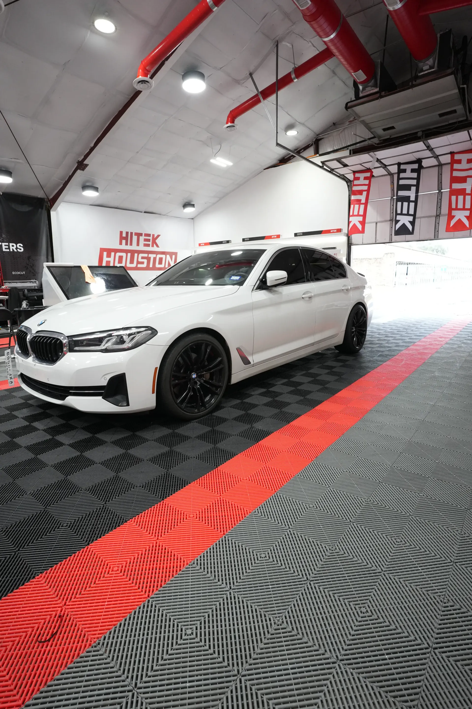 White BMW sedan parked in a garage with black and red flooring and overhead lighting.