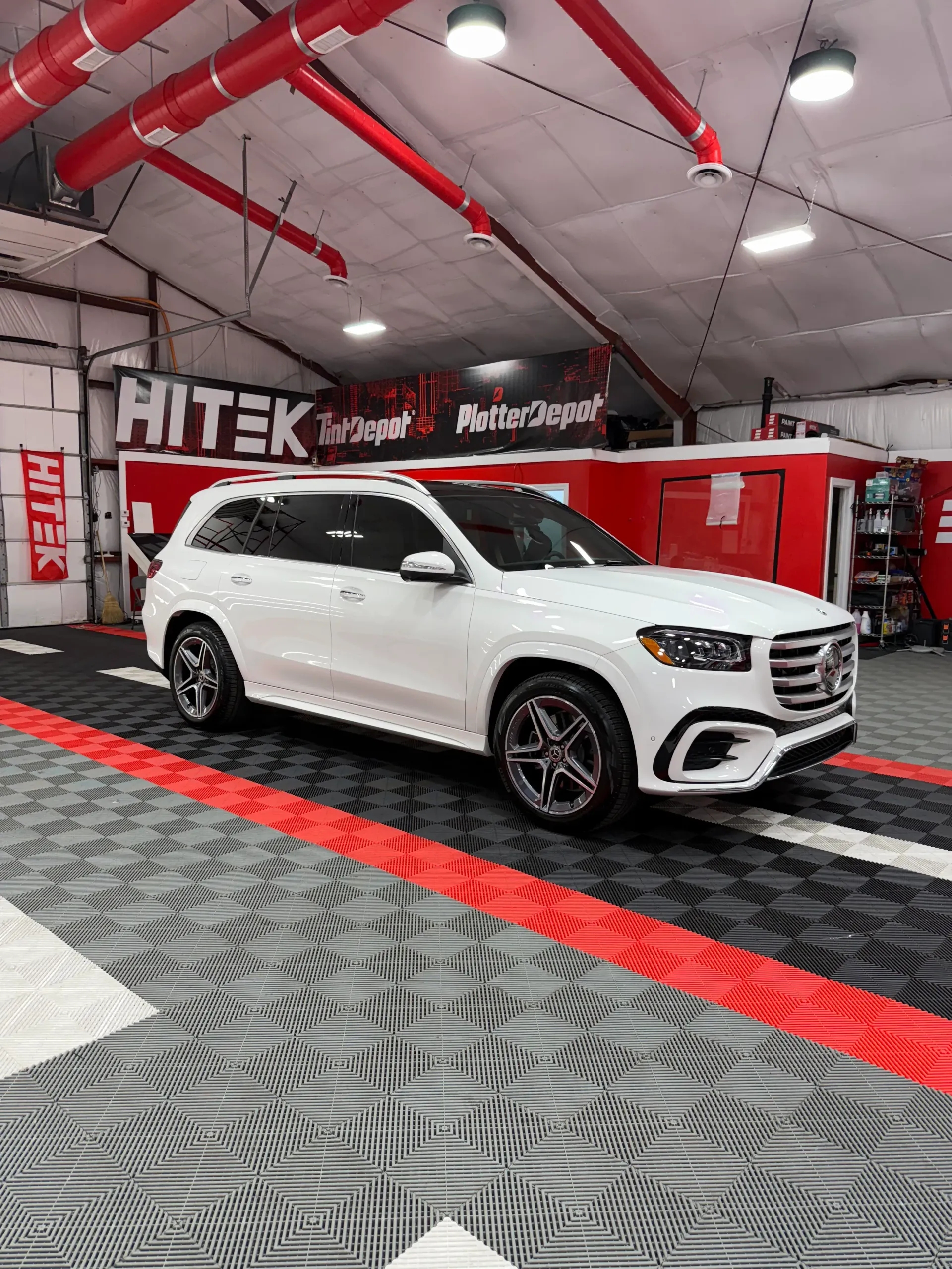 White SUV parked inside a garage with red accents, black and gray checkered floor, and overhead lights.
