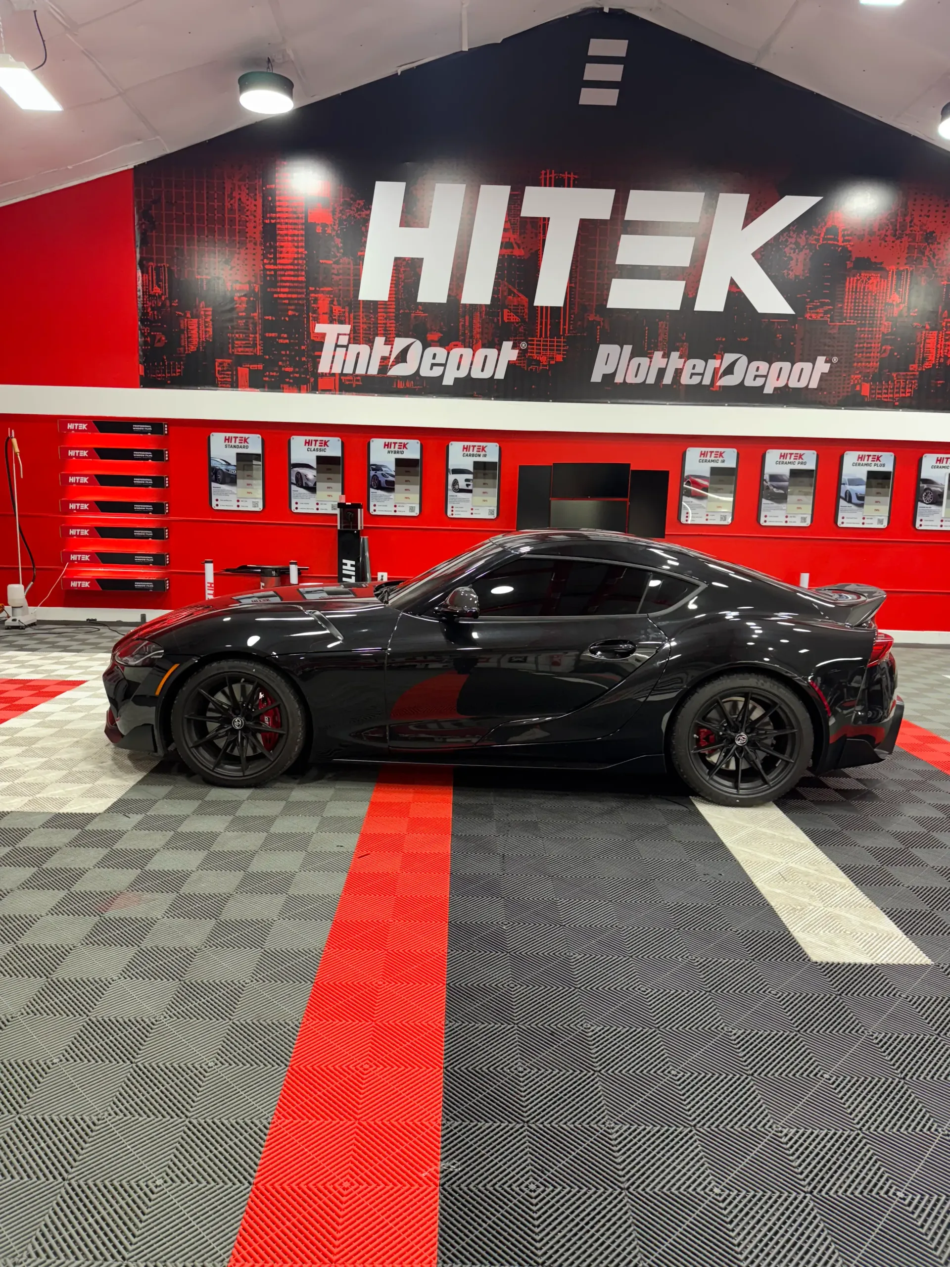 Black sports car parked inside a HiTek Tint Depot. Red and black decor, tiled floor.