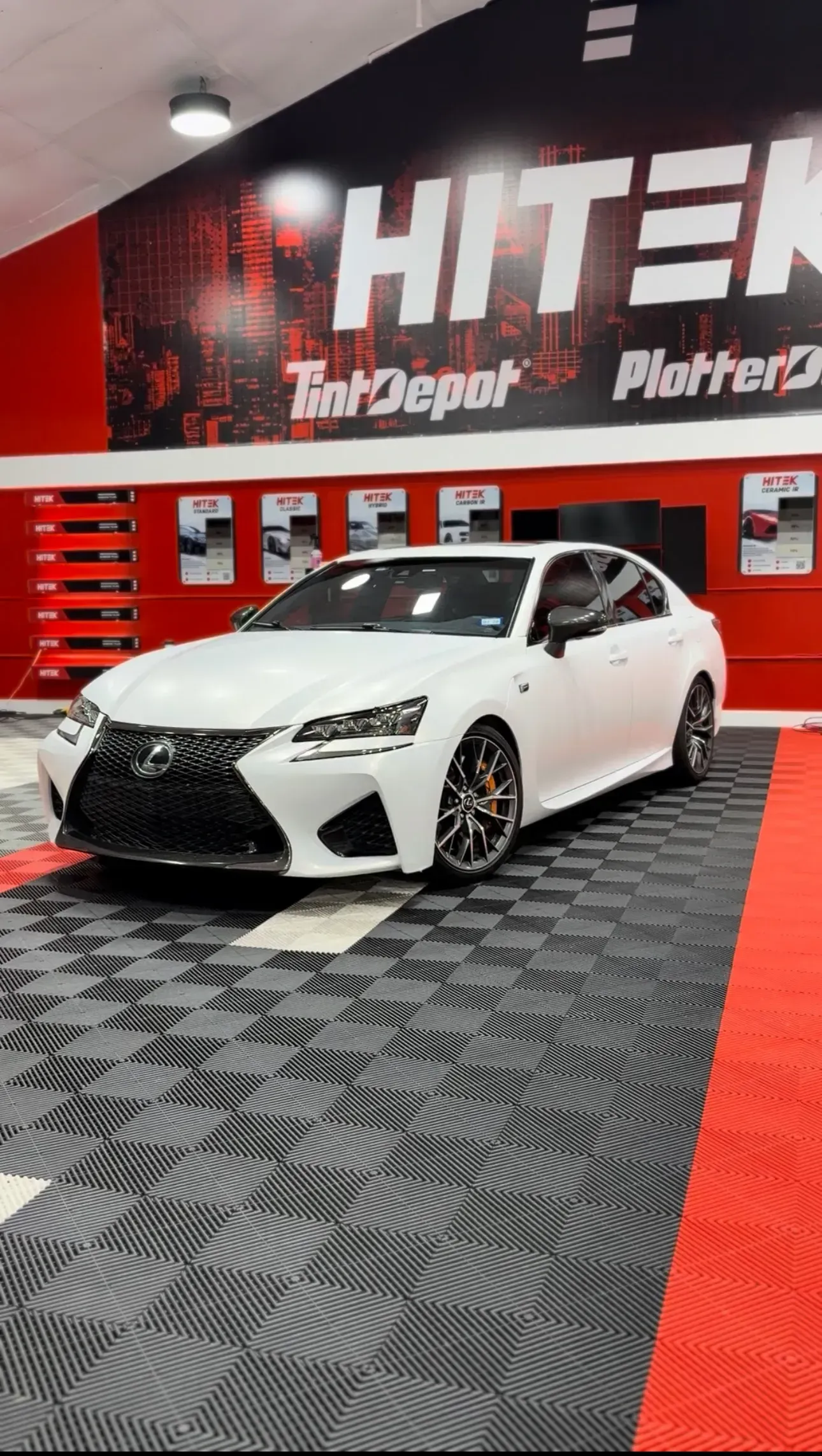 White Lexus GS F car parked inside a facility, with a red and black logo in the background.