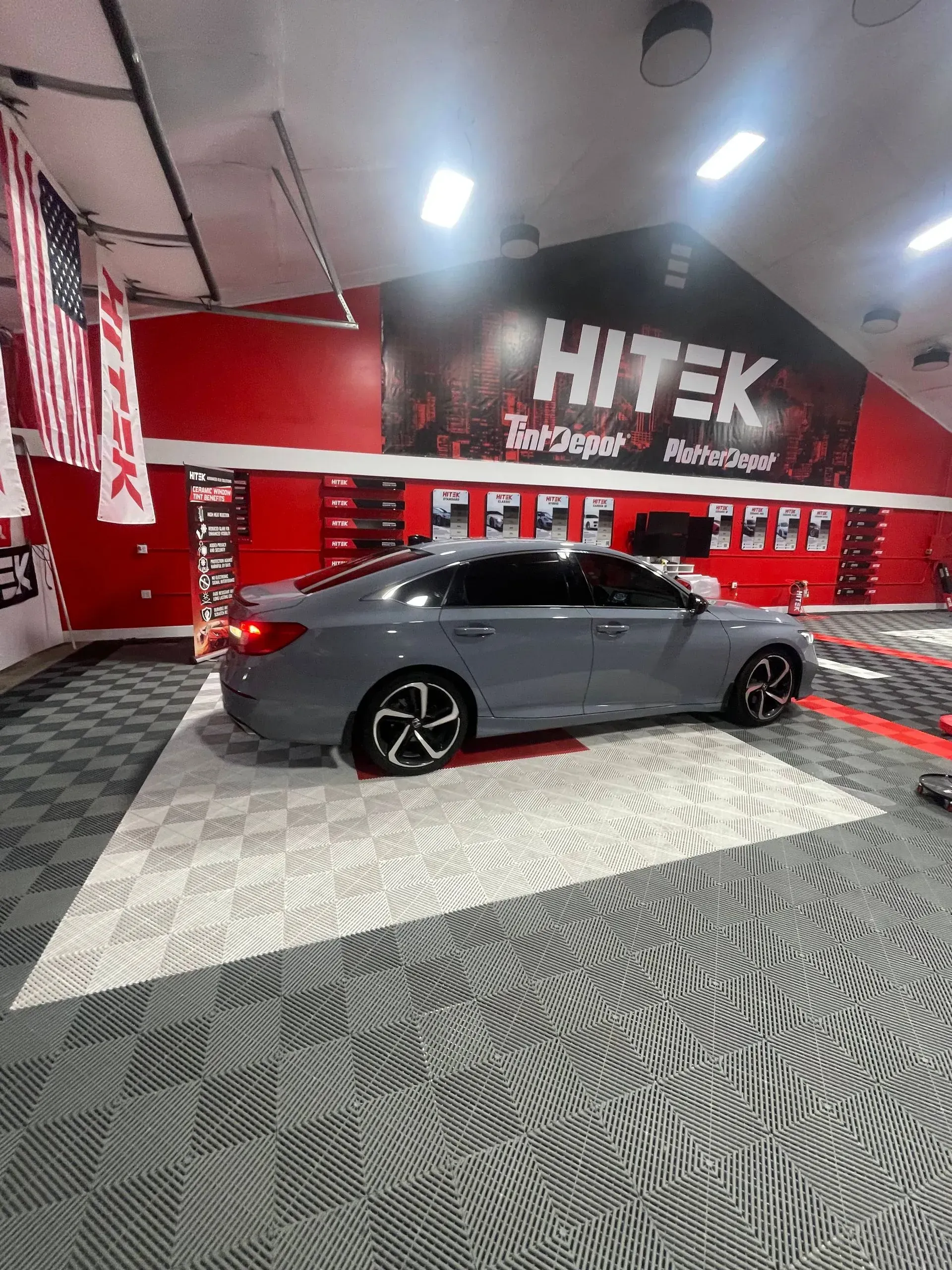 Gray sedan on a checkered floor in a shop with red and black accents, with a banner that reads “HITEK.”