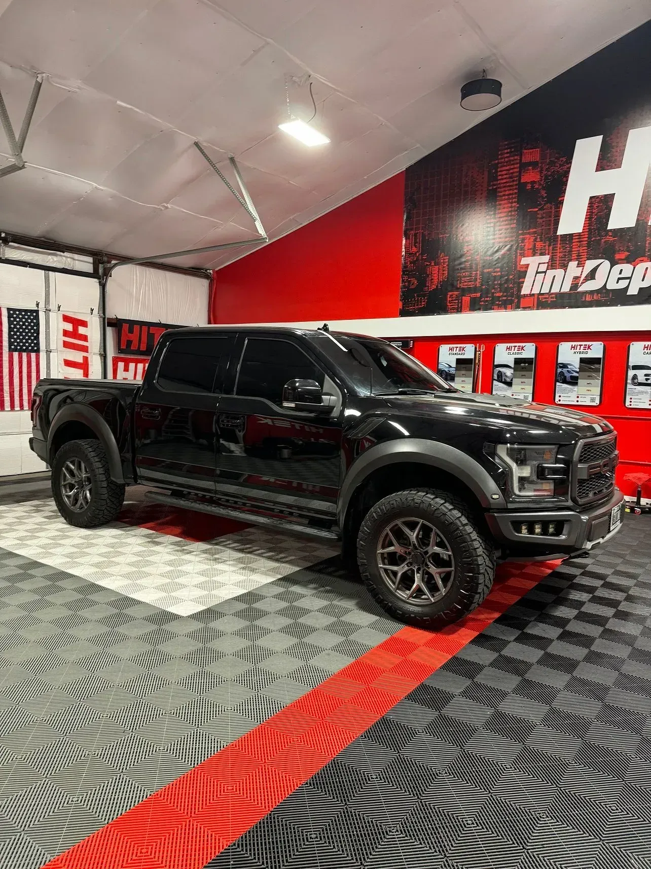 Black Ford Raptor truck inside a garage with red and black flooring and a red wall with logo.