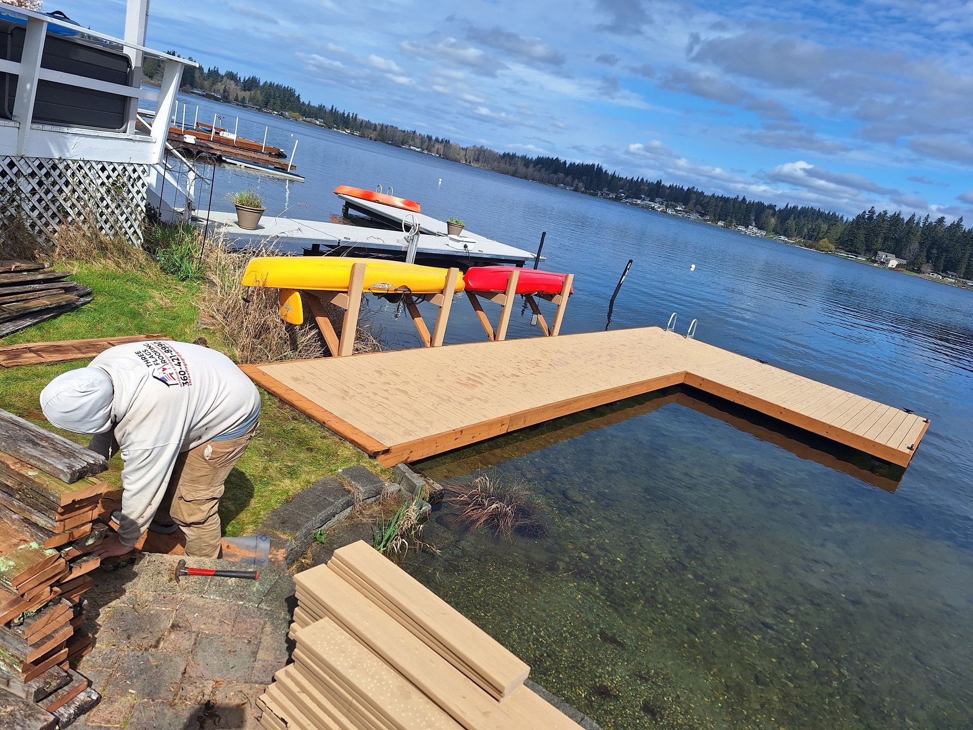 Worker building a wooden dock beside a calm lake, with a red kayak and stacked planks nearby.