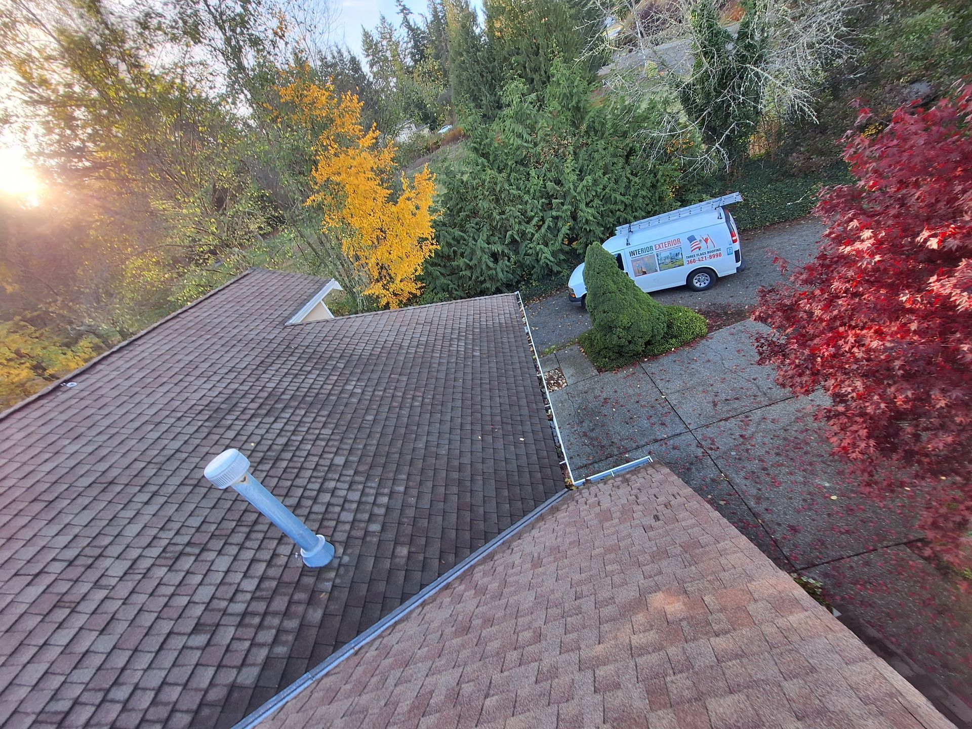 Roof view of a driveway with a white van, autumn trees, and sunlight over shingled rooftops