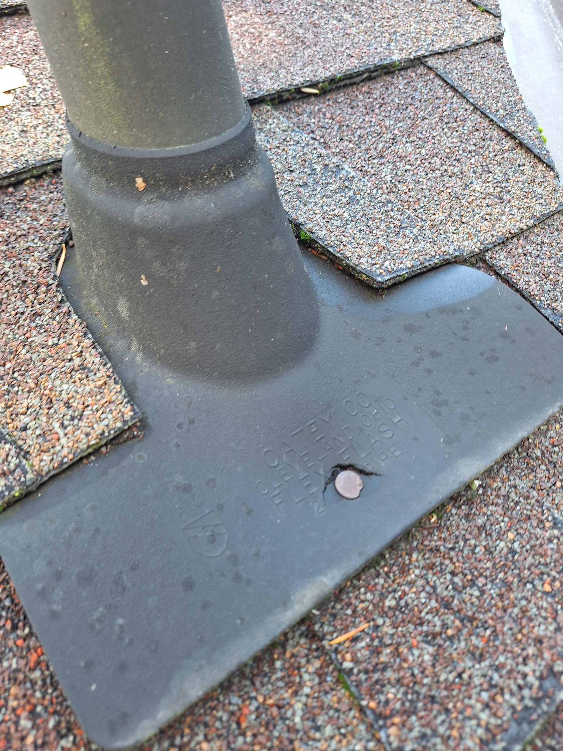 Roof vent pipe base with black flashing on shingled roof, showing a small exposed fastener.