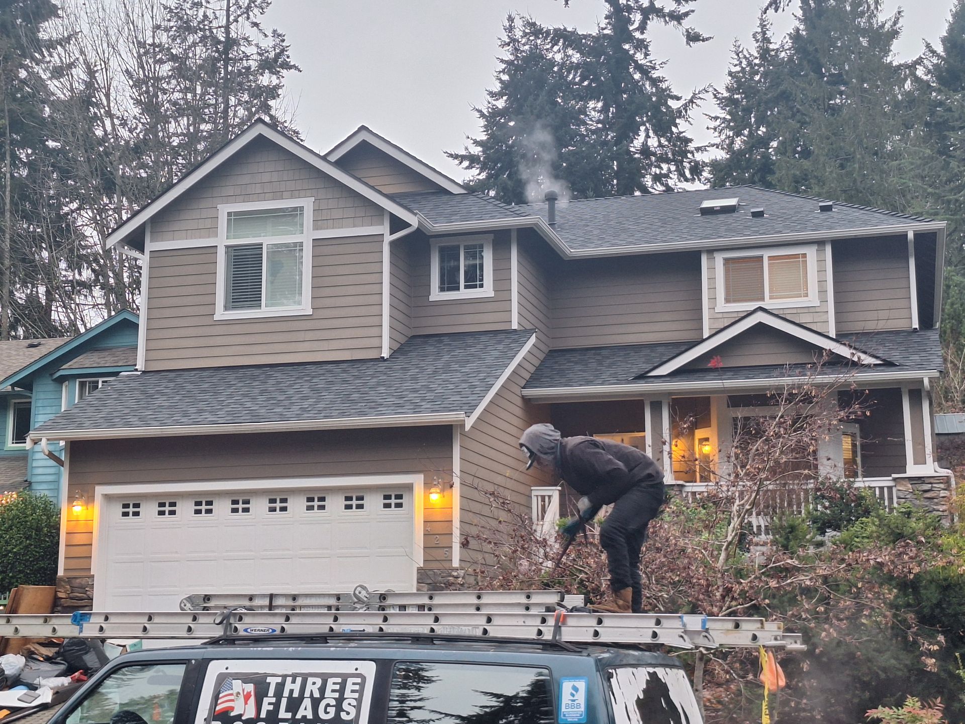 Person working on a roof of a suburban house with a garage, surrounded by trees.