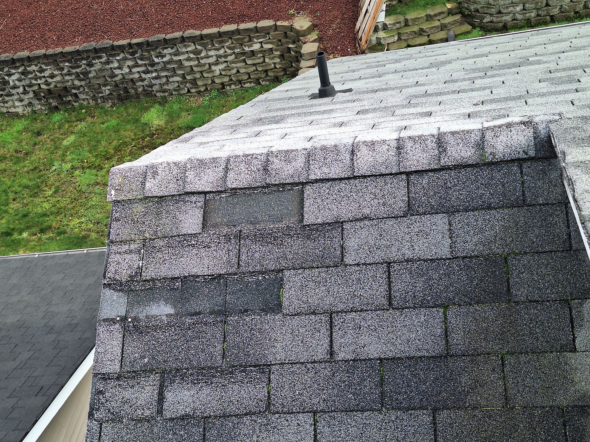 Close-up of a gray shingled roof with a black vent pipe beside a brick wall and grass.
