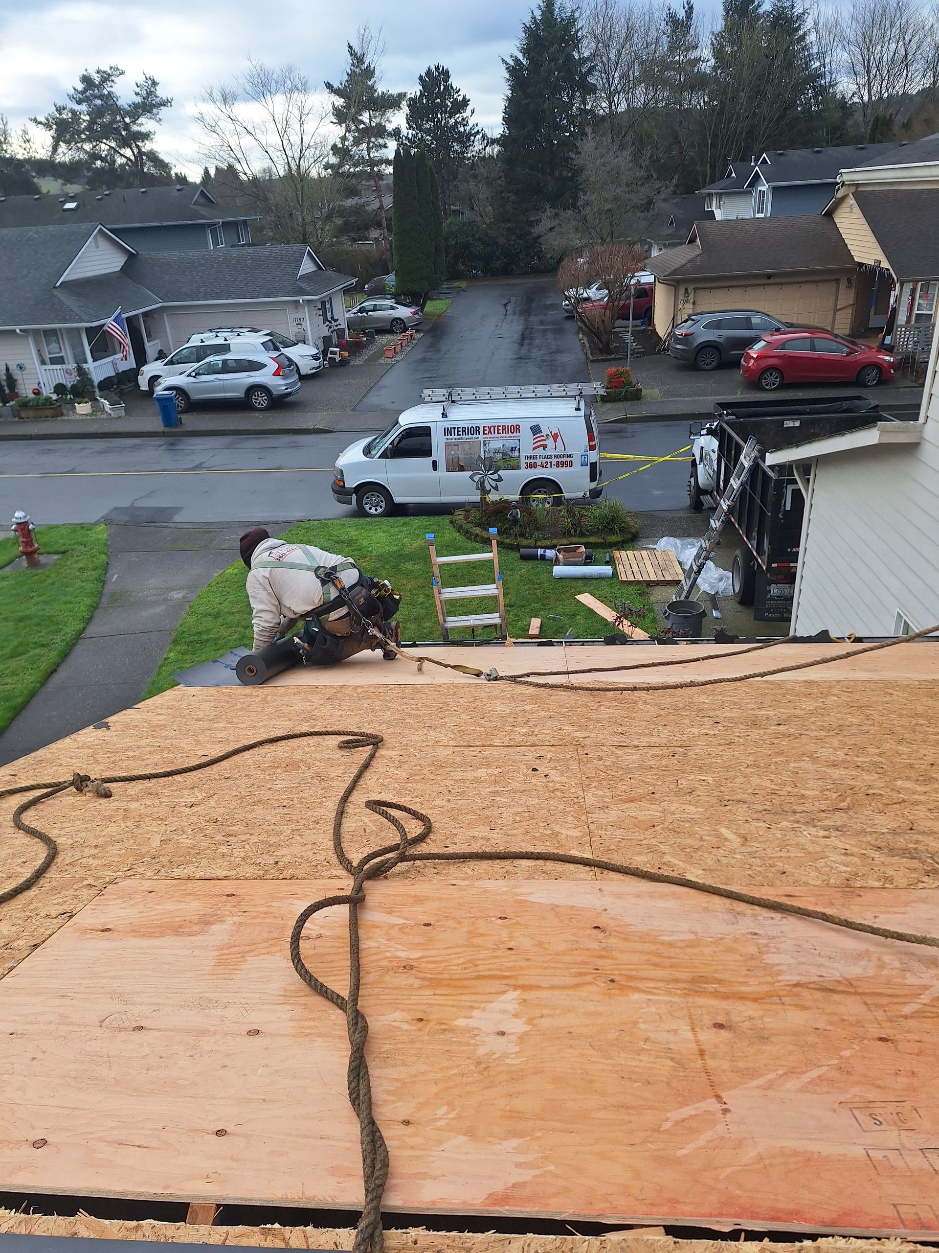 Roofing work on a house with plywood underlayment, workers, and trucks on a suburban street