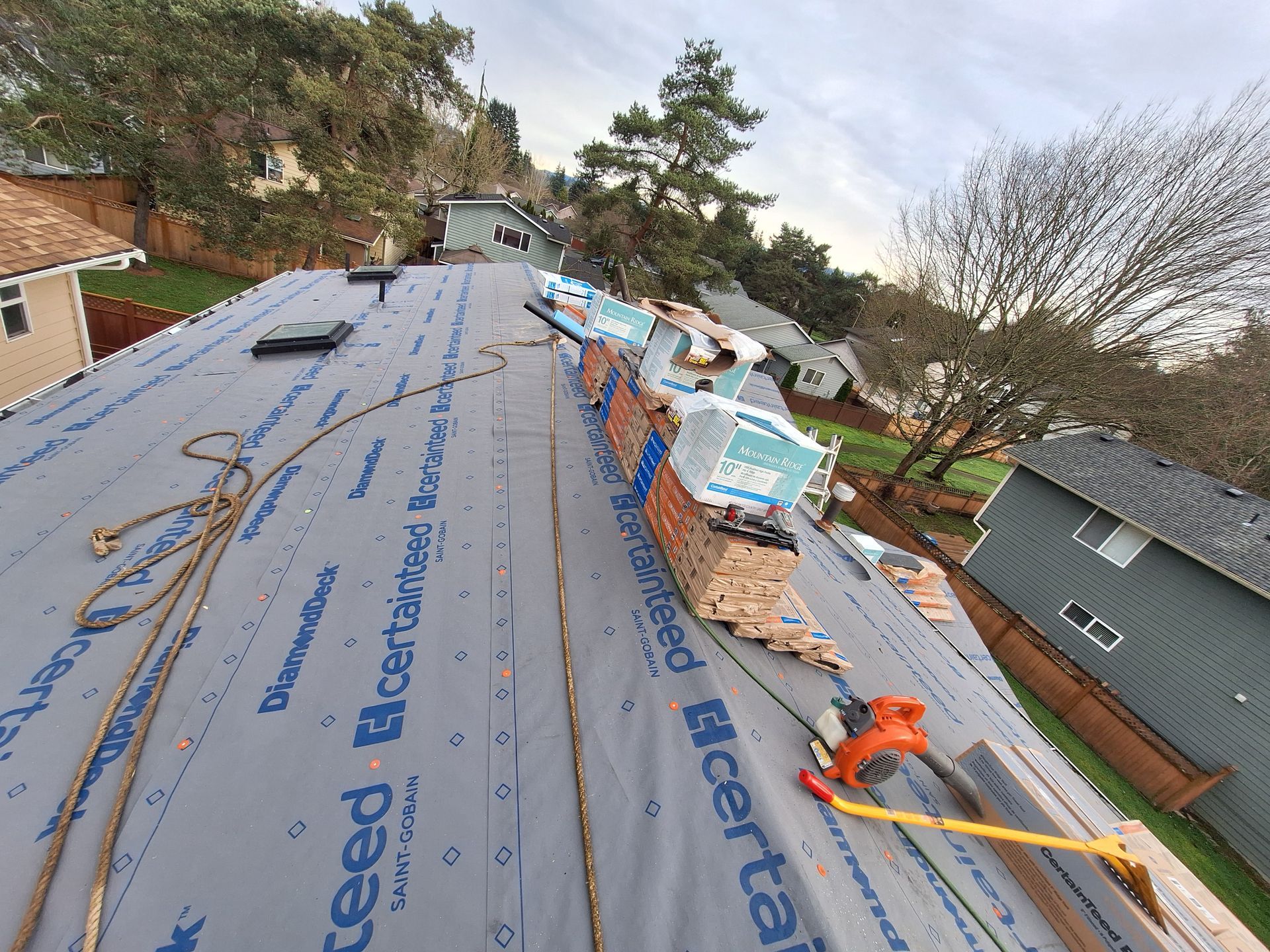 Roof under repair with blue underlayment, tools, and workers along a suburban house roofline