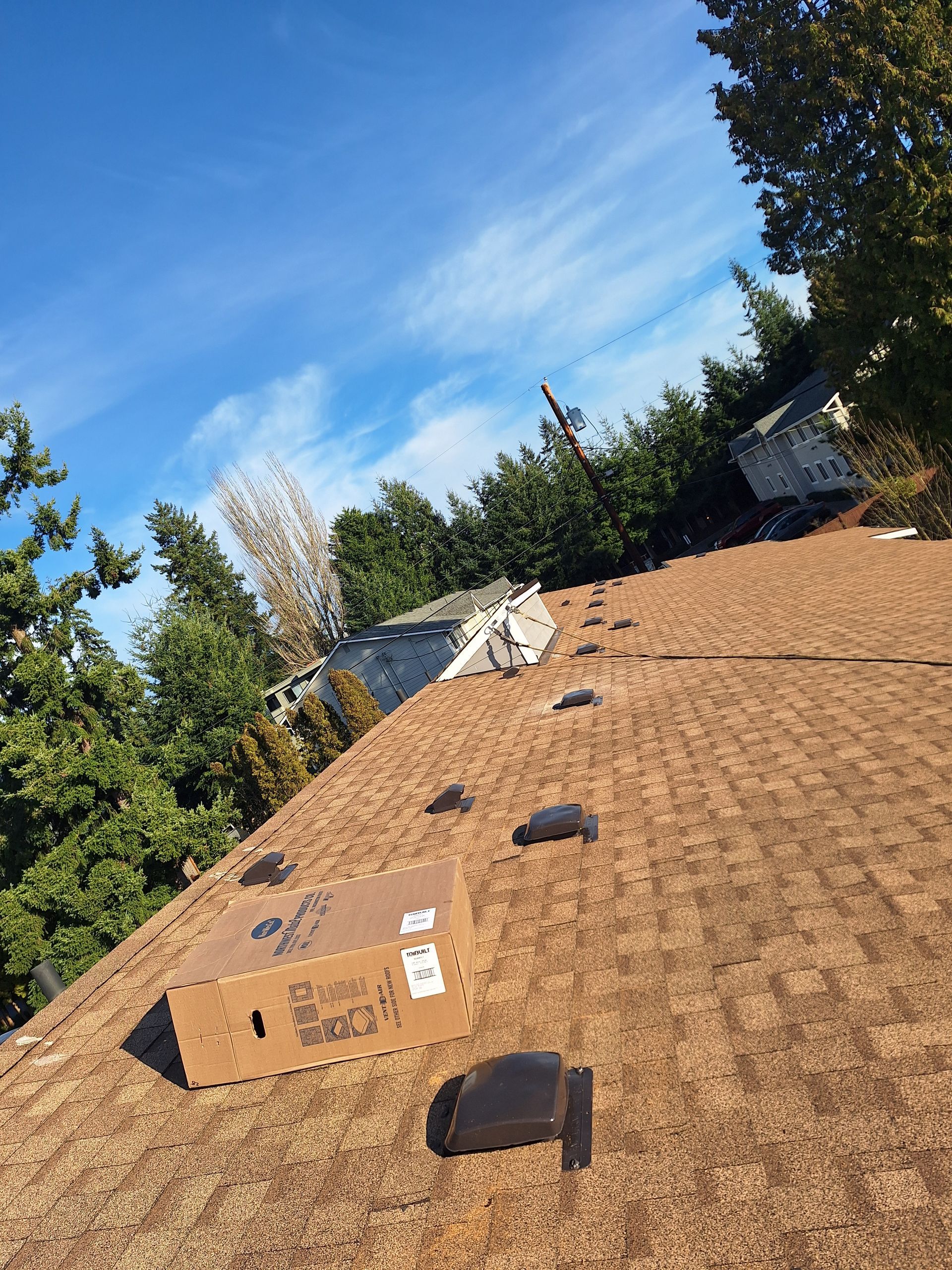 Roof under construction with brown shingles, vents, and a cardboard box under a blue sky.