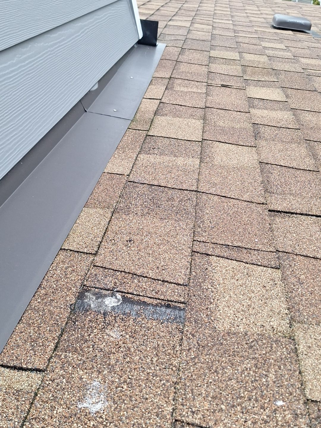 Close-up of a shingled roof beside gray siding, showing a damaged patch and white debris.