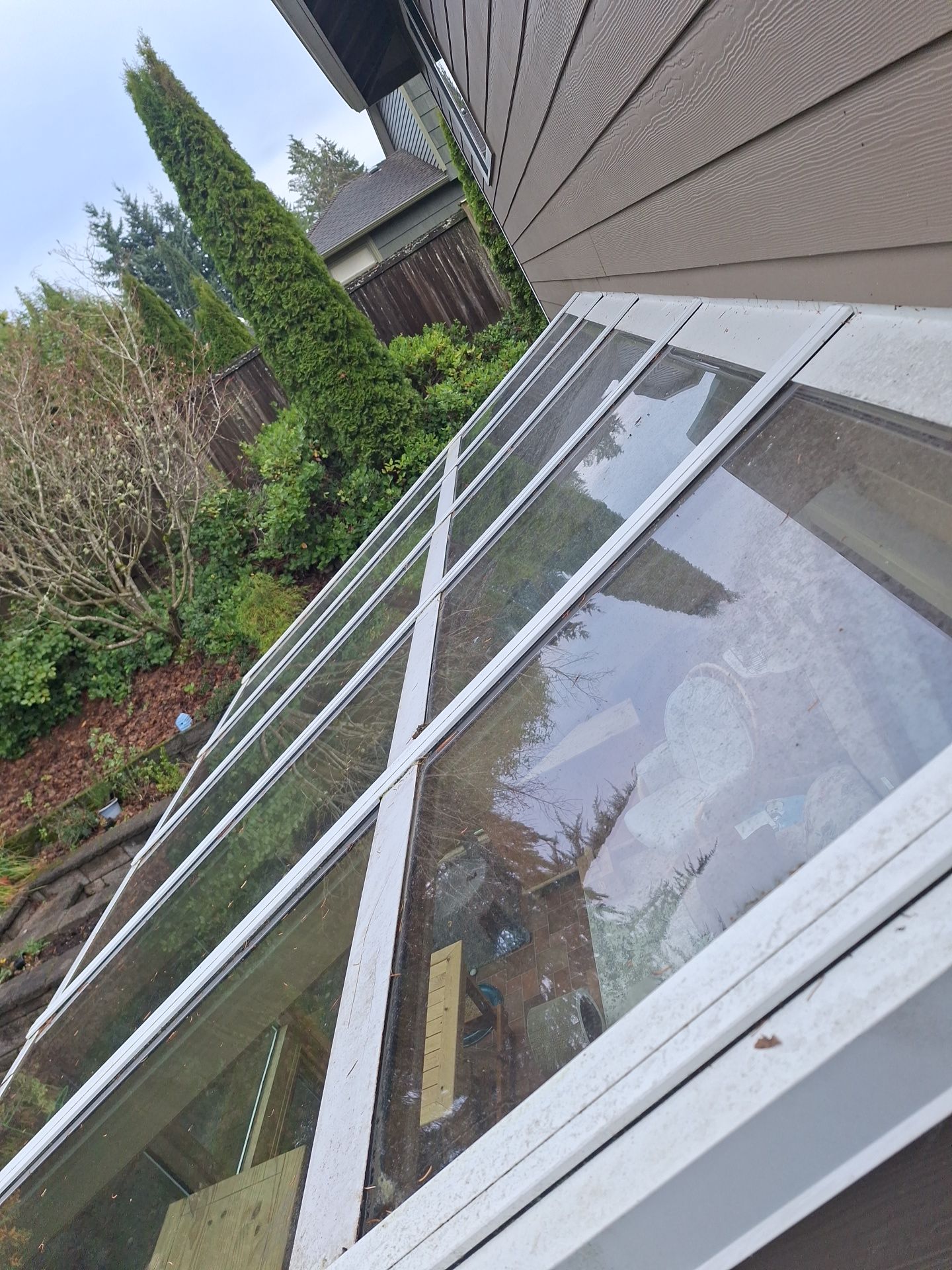 Broken glass roof windows along a house exterior beside a backyard garden.