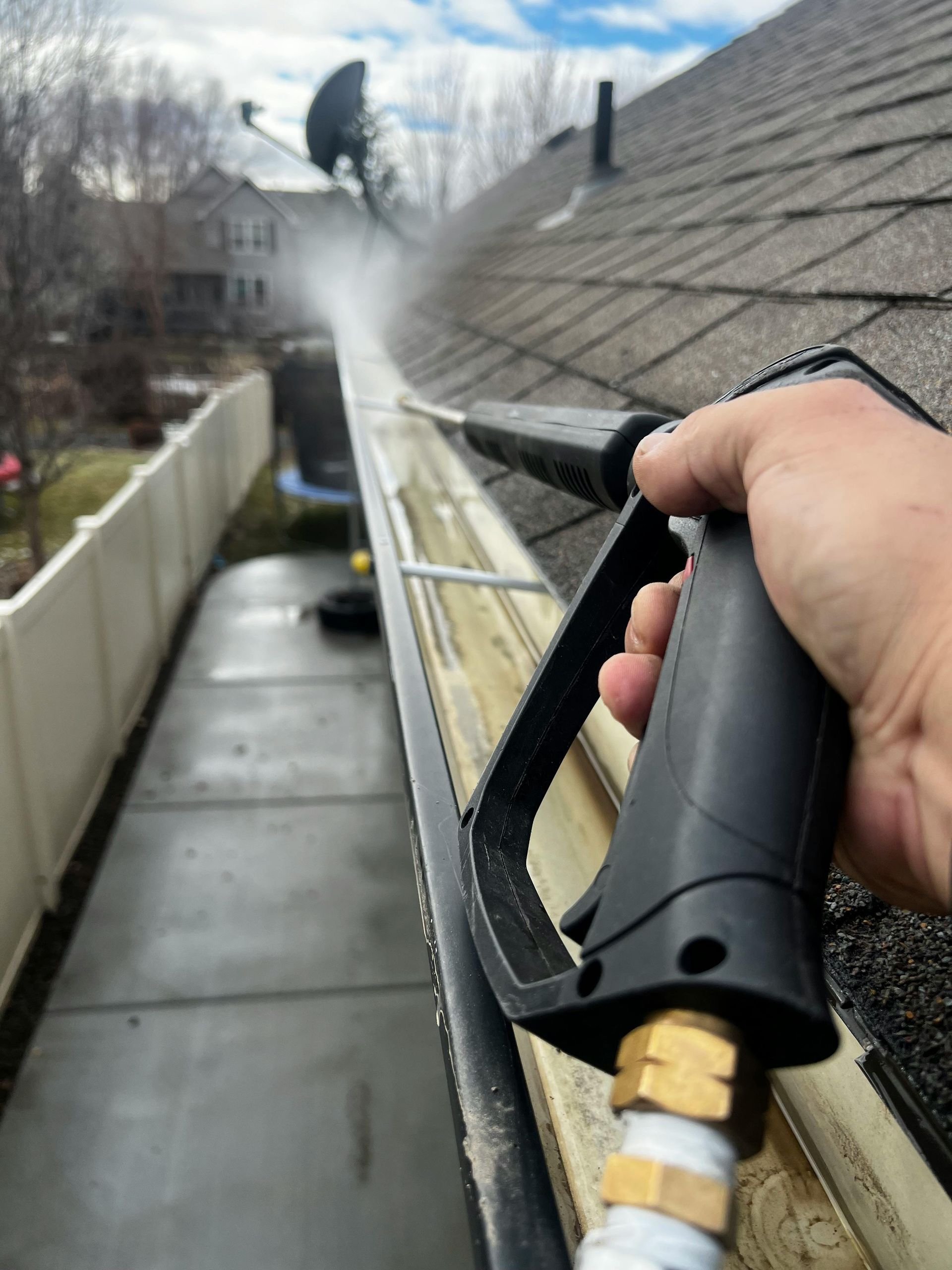 Hand holding pressure washer spraying water along a roof gutter beside a house