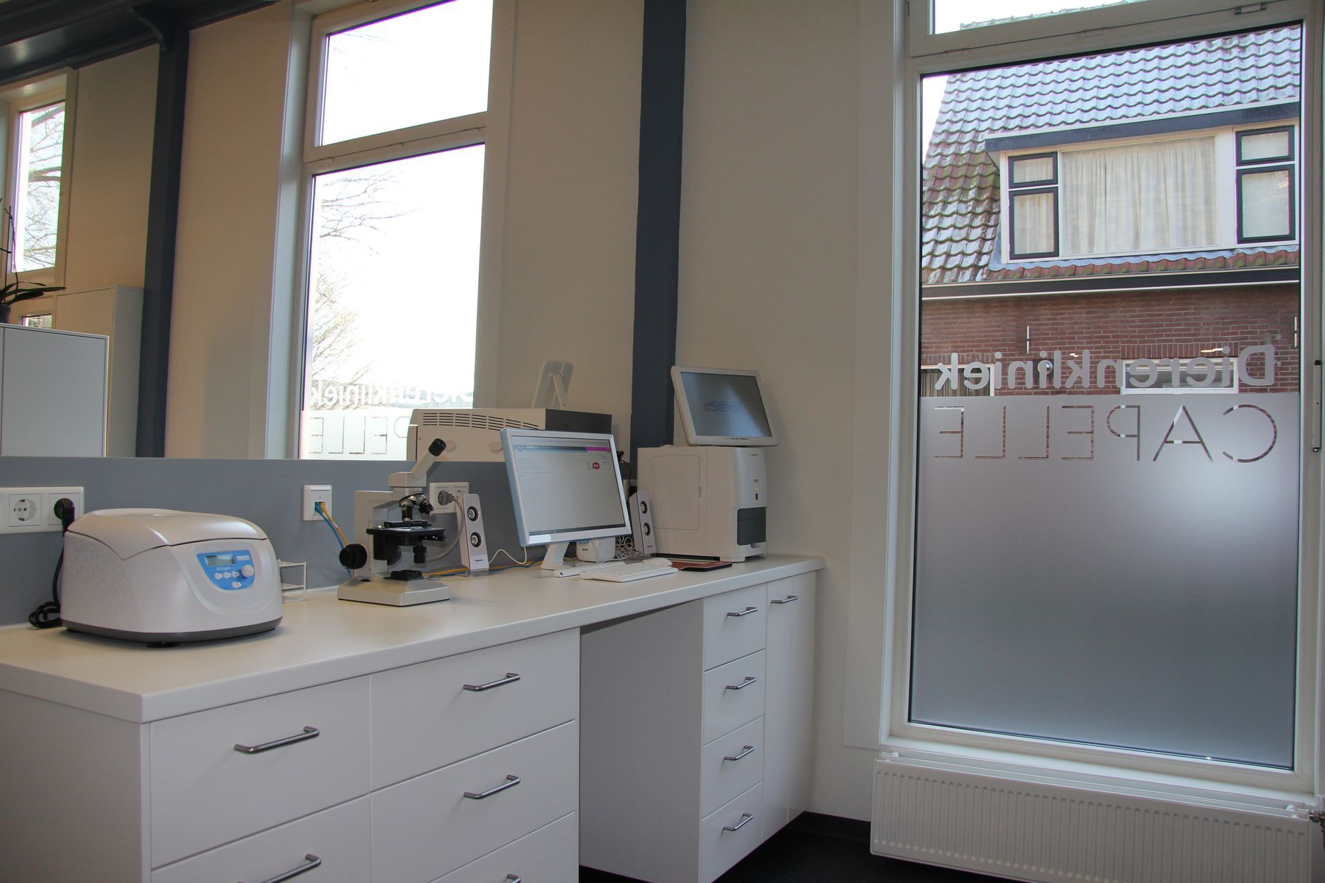 Licht kantoor met witte kasten, een microscoop en een monitor op een balie bij grote ramen.