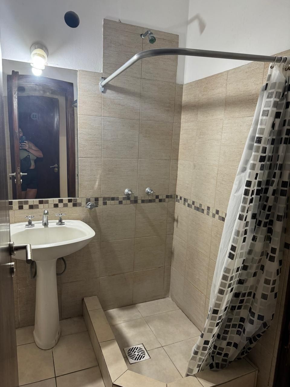 Un baño con azulejos de color beige, con un lavabo de pedestal a la izquierda y una zona de ducha