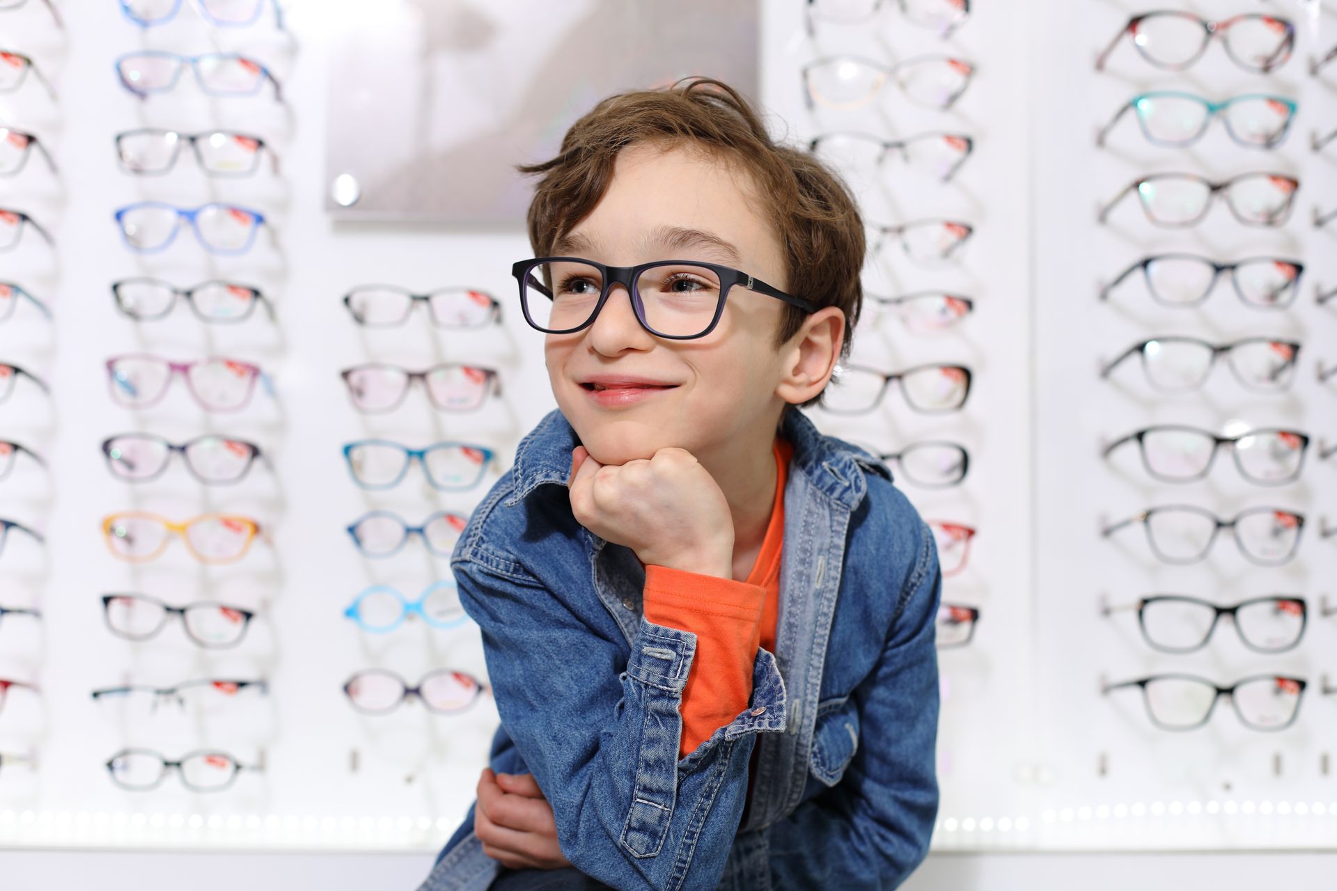 Person in a blue jacket and orange shirt posing in an eyeglasses store, smiling in front of wall displays.
